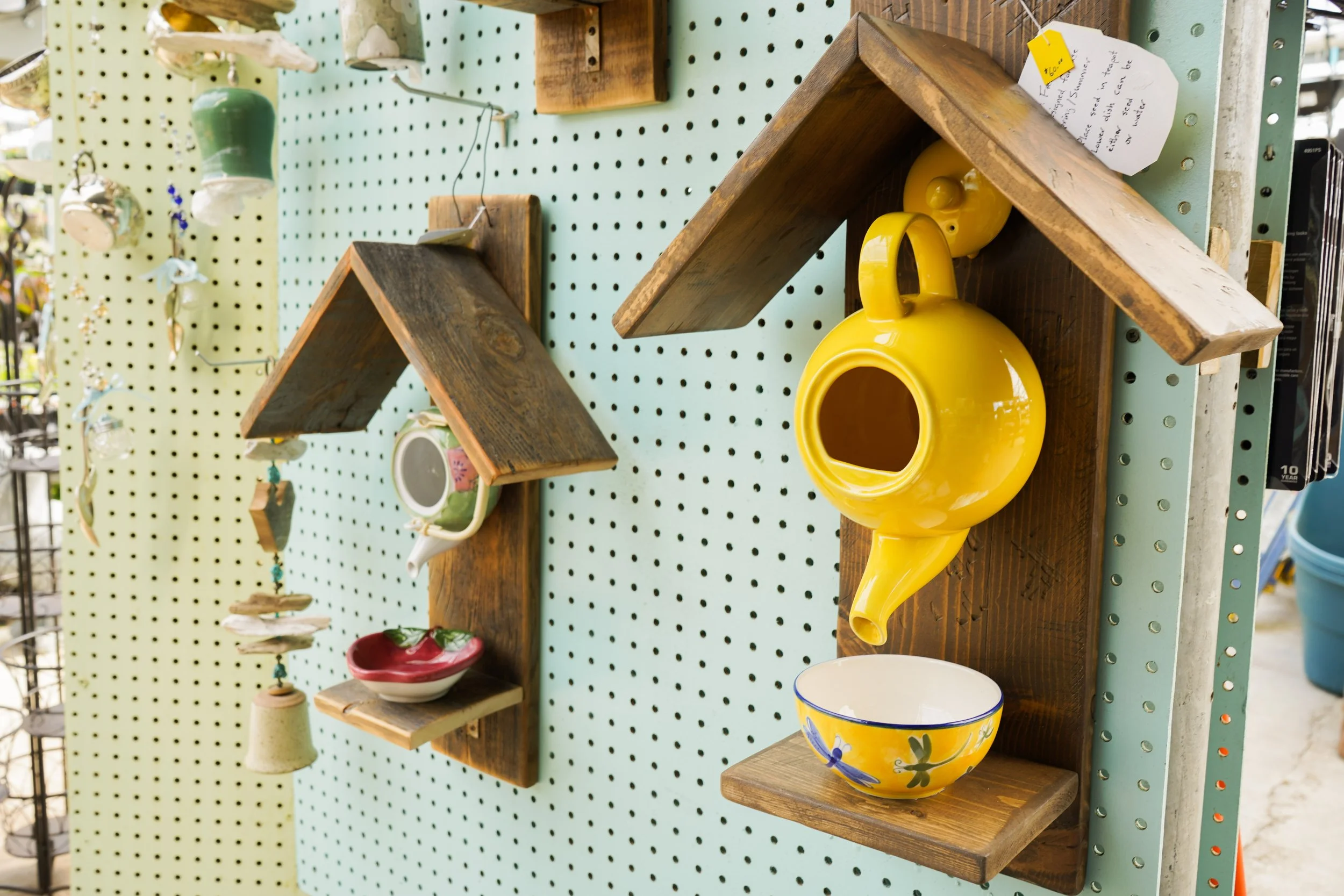 Display of various ceramic and wooden decorative items on a pegboard, including a yellow ceramic teapot, colorful bowls, and other small ornaments.