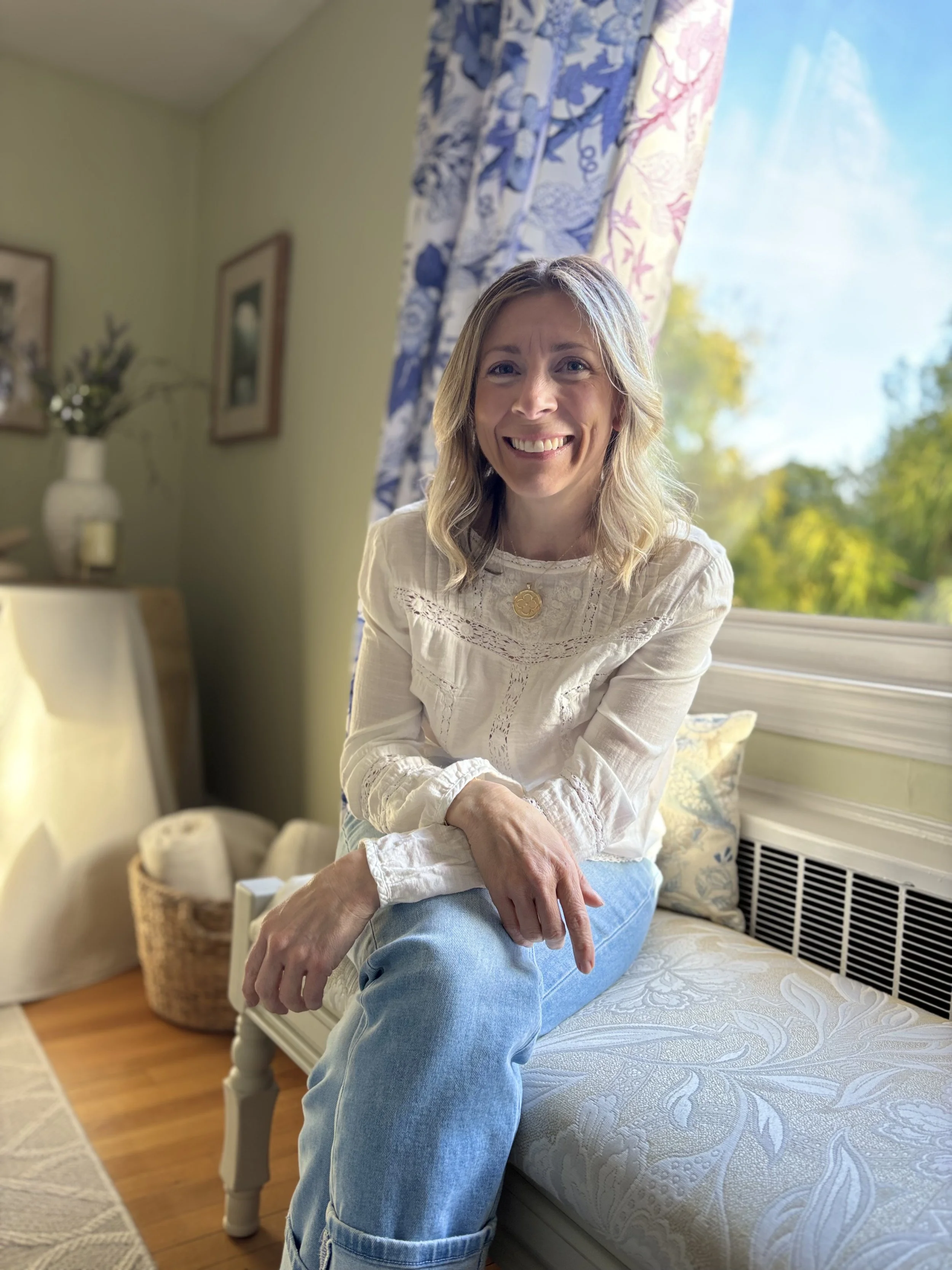 A smiling woman with blonde hair in a white embroidered blouse and blue jeans sitting on the edge of a bed in a room with green walls and floral curtains, with sunlight coming through a window behind her.