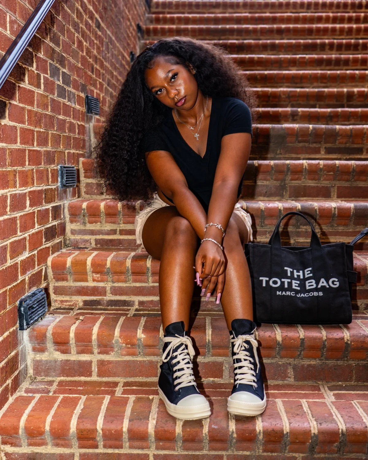 Young woman sitting on brick stairs with a black tote bag, wearing a black t-shirt, beige shorts, and black and white sneakers.