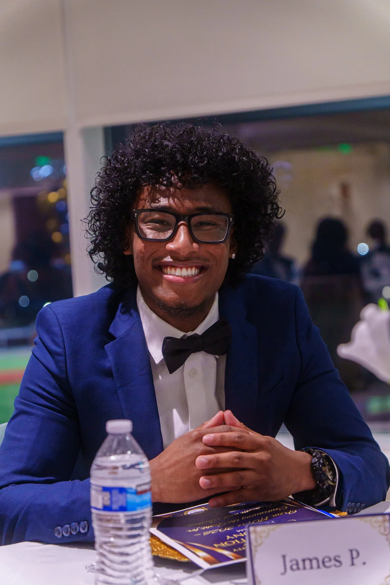 Young man with curly black hair, glasses, and a big smile, wearing a blue suit, white shirt, and black bow tie, sitting at a table with a water bottle, a name tag labeled 'James P.', and a program or menu, in a well-lit indoor setting with people in 