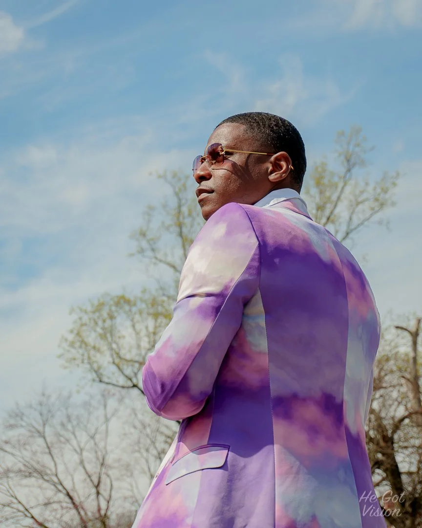 A man wearing a purple and white tie-dye blazer and sunglasses is standing outdoors against a blue sky with some trees in the background.