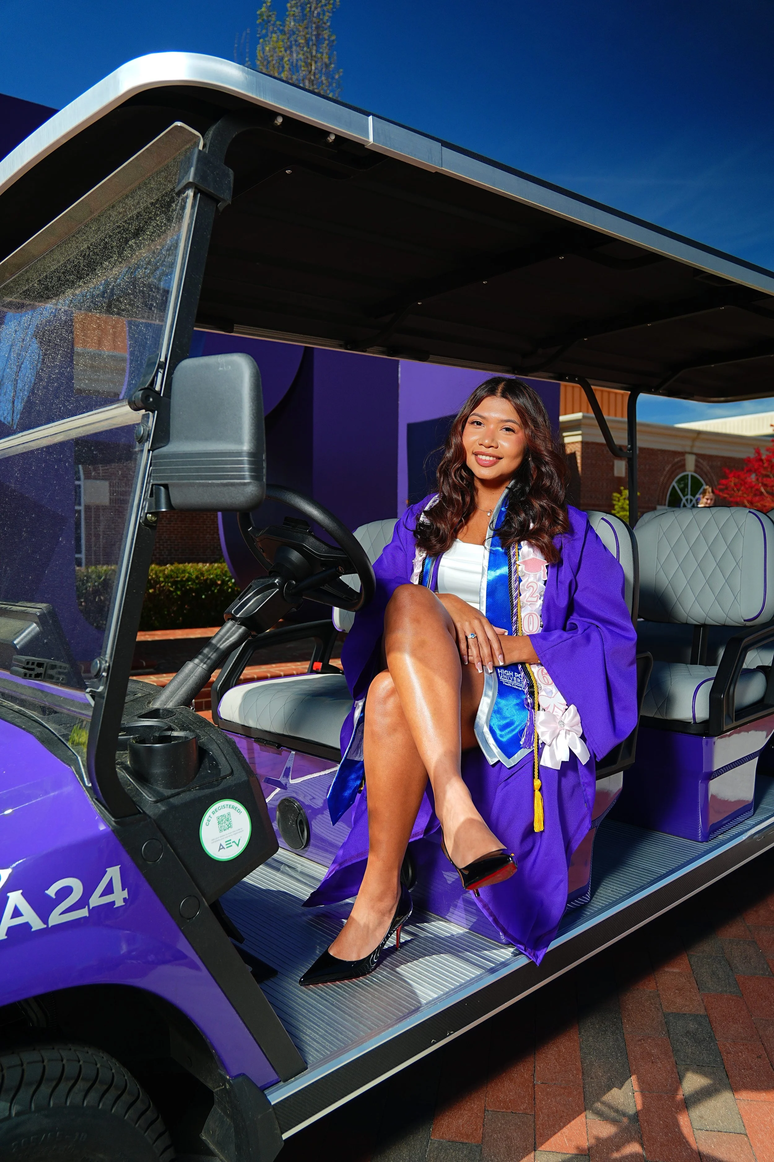 A young woman in a graduation gown and sash sitting inside a purple golf cart with a smile.