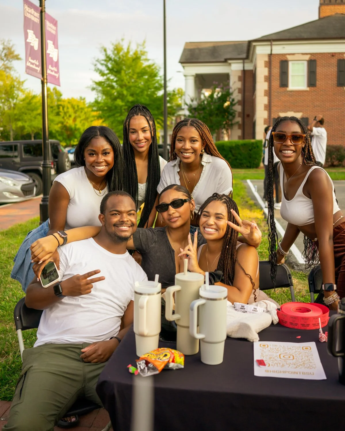 Group of seven young adults smiling and posing outdoors at a gathering, sitting and standing around a table with drinks and snacks.