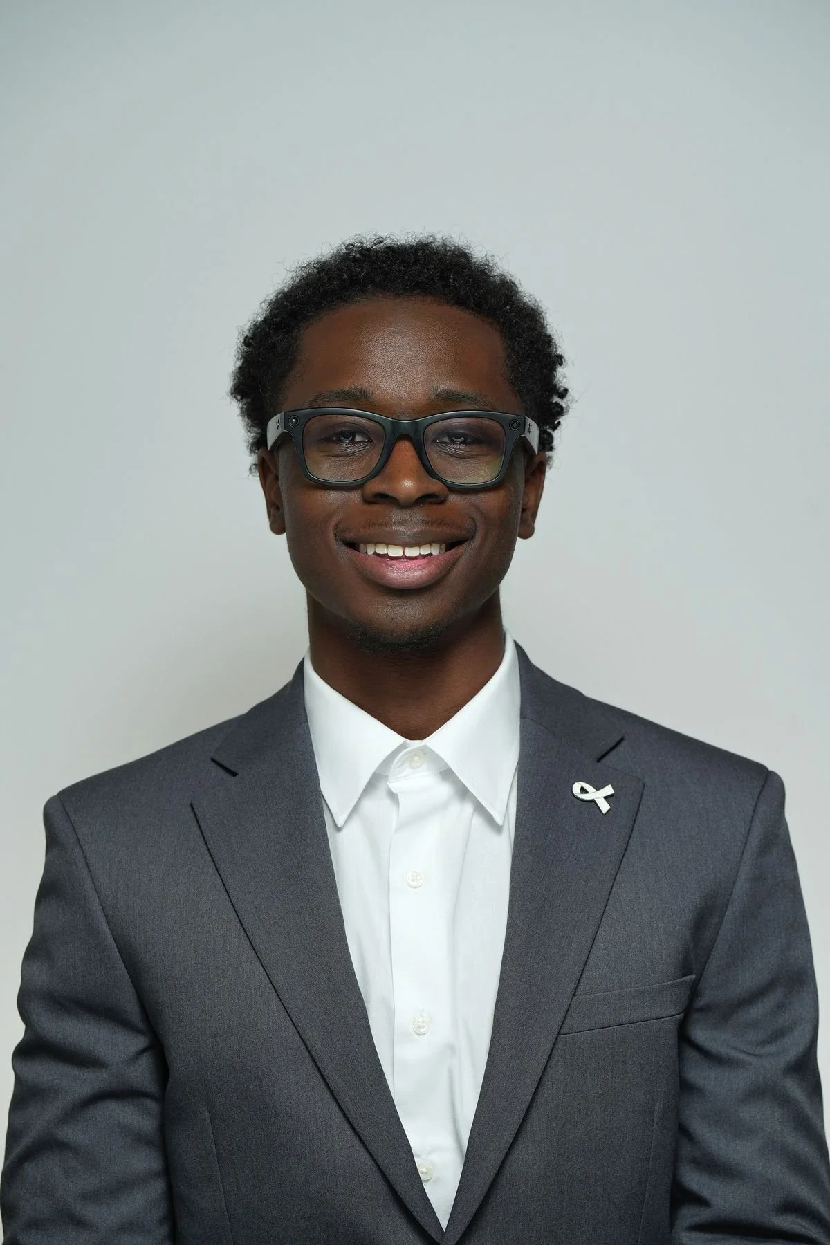 A smiling man wearing a gray suit, white shirt, and glasses, with a disability awareness pin on his lapel, standing against a plain gray background.