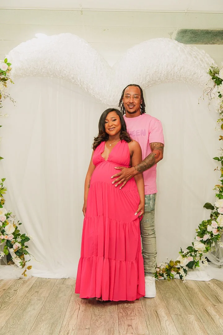 A pregnant woman in a pink dress and a man in a pink T-shirt and jeans standing together in front of a white backdrop with large white heart-shaped decoration and floral arrangements, celebrating a special occasion.