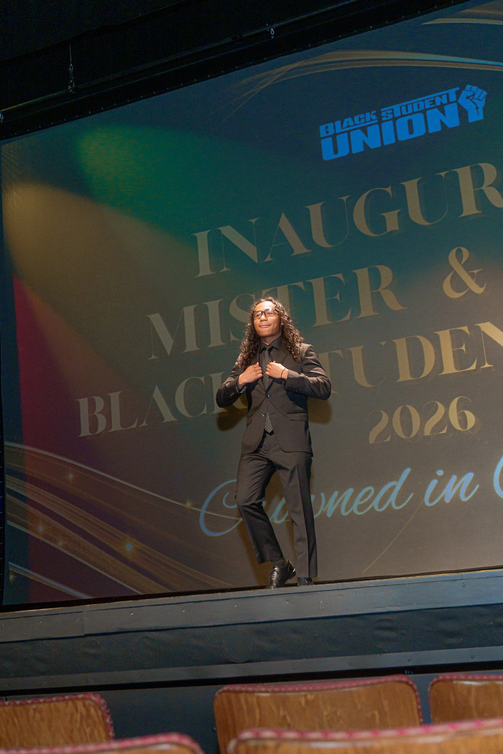 A woman dressed in a black suit on stage at the Black Student Union's Miss and Mister event, with a large screen in the background displaying event information.