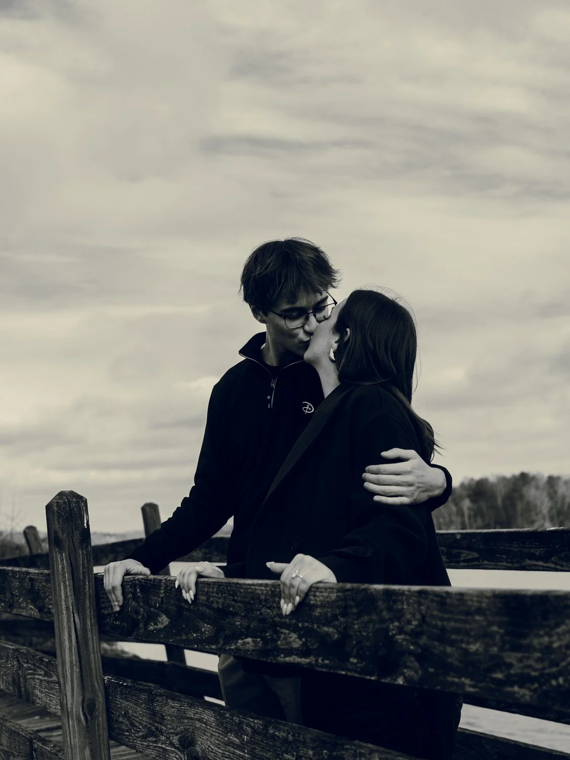 A black-and-white photo of a couple kissing on a wooden bridge outdoors with trees and cloudy sky in the background.