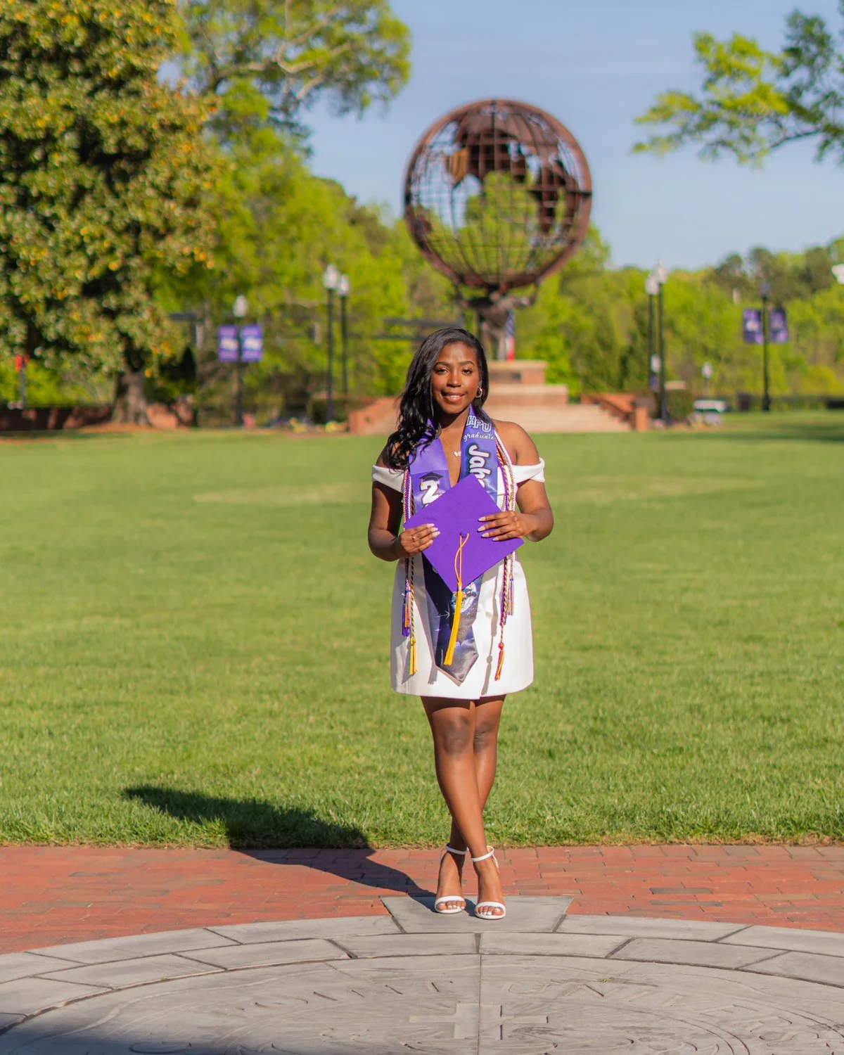 A young woman in a white graduation dress holding a purple diploma cover, standing outdoors on a brick platform, with a monument of a globe in the background and green trees around on a bright day.
