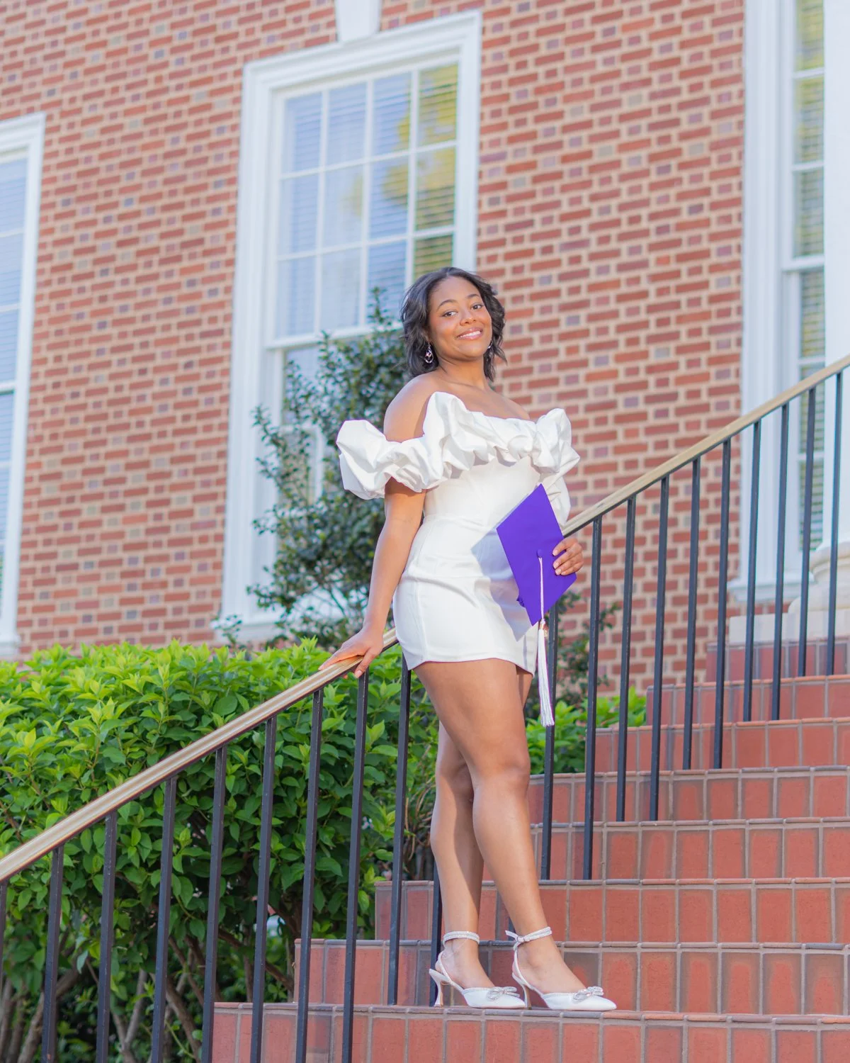 Young woman in white dress standing on brick stairs outside a brick building, holding a purple diploma, smiling.