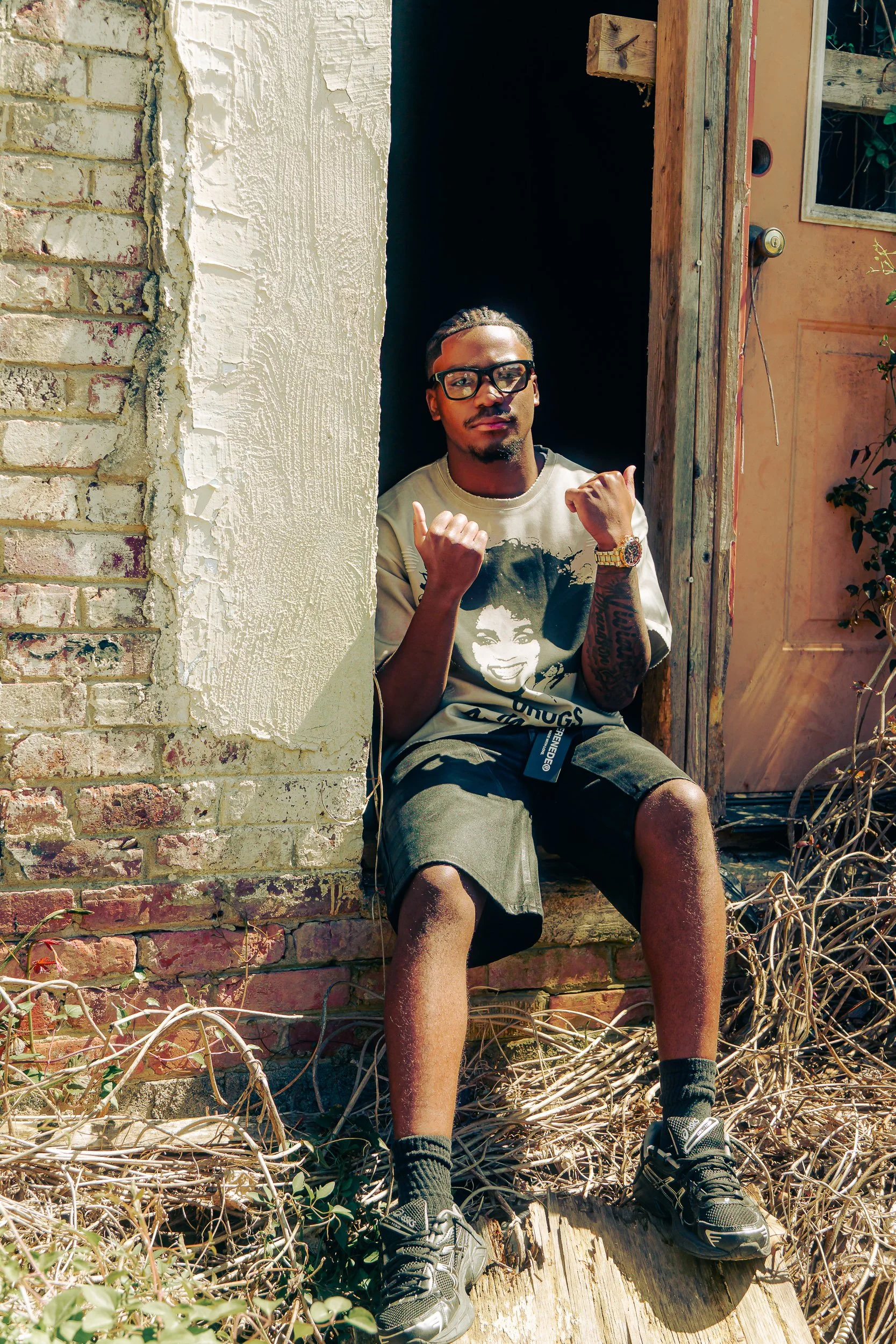 A young man with glasses and a graphic t-shirt sitting on a step outside a building, making a peace sign with both hands. There are dried plants around his feet, and the building has a brick and stucco wall with a wooden door.