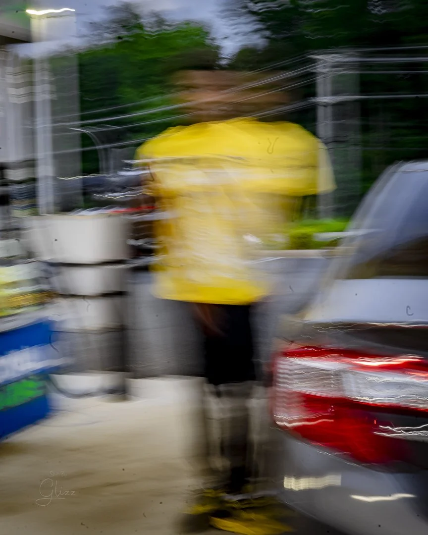 Blurred image of a person standing outdoors near a vehicle, wearing a yellow shirt, with trees in the background.