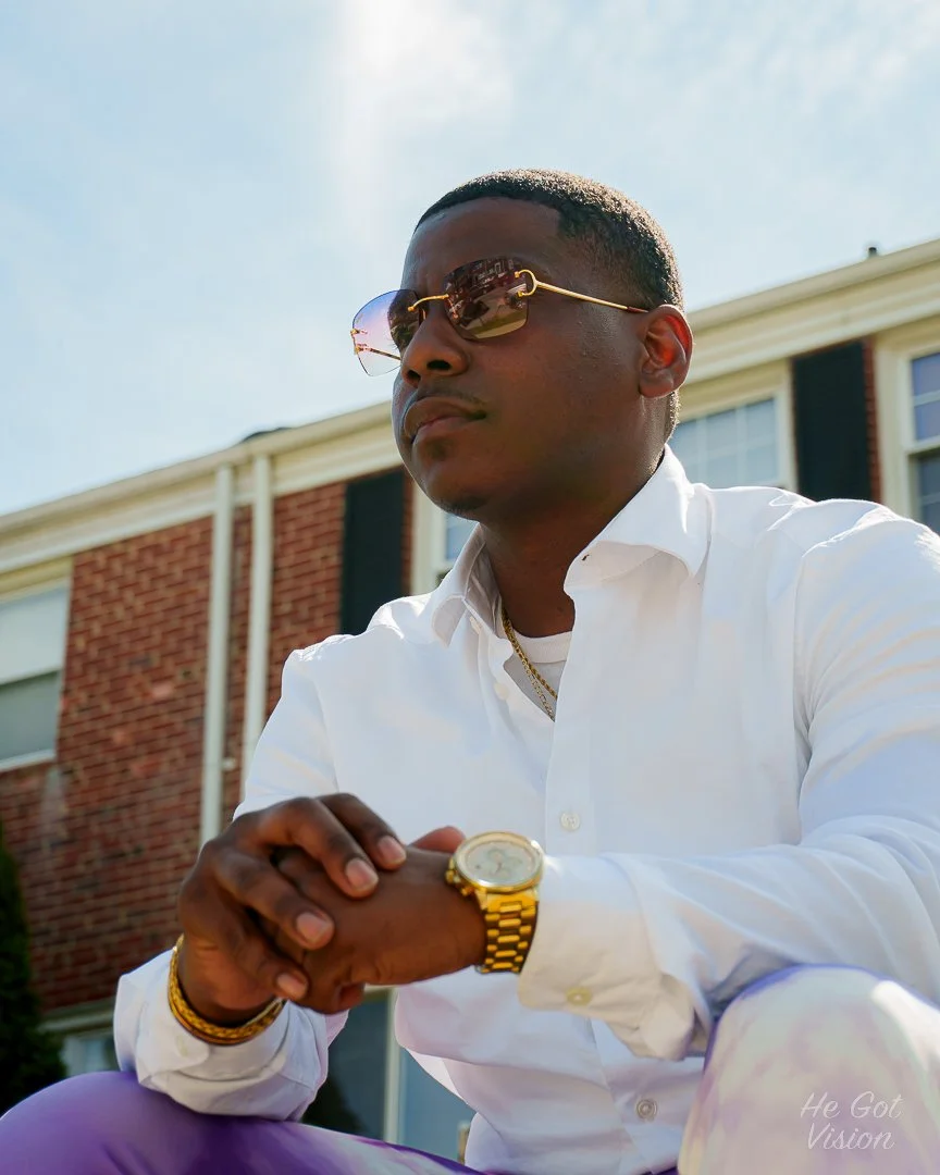 A young man wearing sunglasses, a white shirt, and gold jewelry, sitting outdoors in front of a house with red brick walls. He has a gold watch and bracelet, and a gold chain, with his hands clasped together.