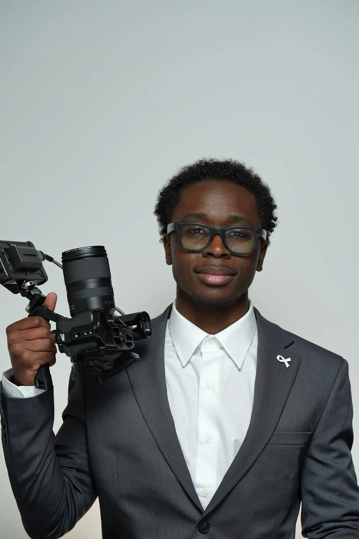 A man wearing a gray suit, white shirt, and glasses holding a camera against a plain background.