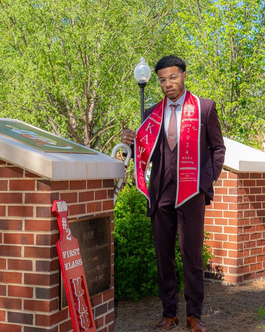A young man in a dark suit and glasses standing outdoors with a red graduation stole and a sash, holding a white cane, near a brick monument with a plaque and a red foam sign that says "110 #2 FIRST CLASS KAPPA". Green trees and a lamp post are in th