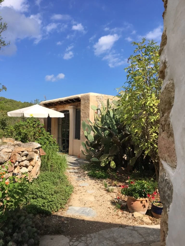 Ein kleines, erdiges Cottage mit einer terrakottafarbenen Fassade und terrassenförmigen Fenstern inmitten eines Garten mit verschiedenen Pflanzen auf Ibiza.