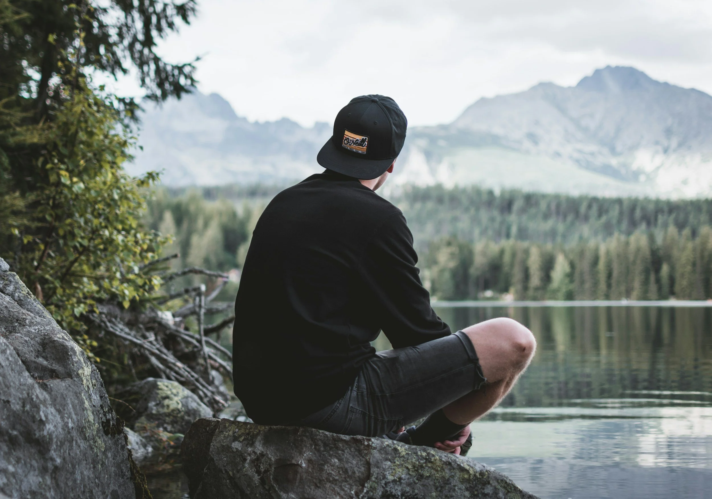 A person wearing a black hat and black shirt sitting on a rock by a lake, looking at mountains and a forest in the distance.