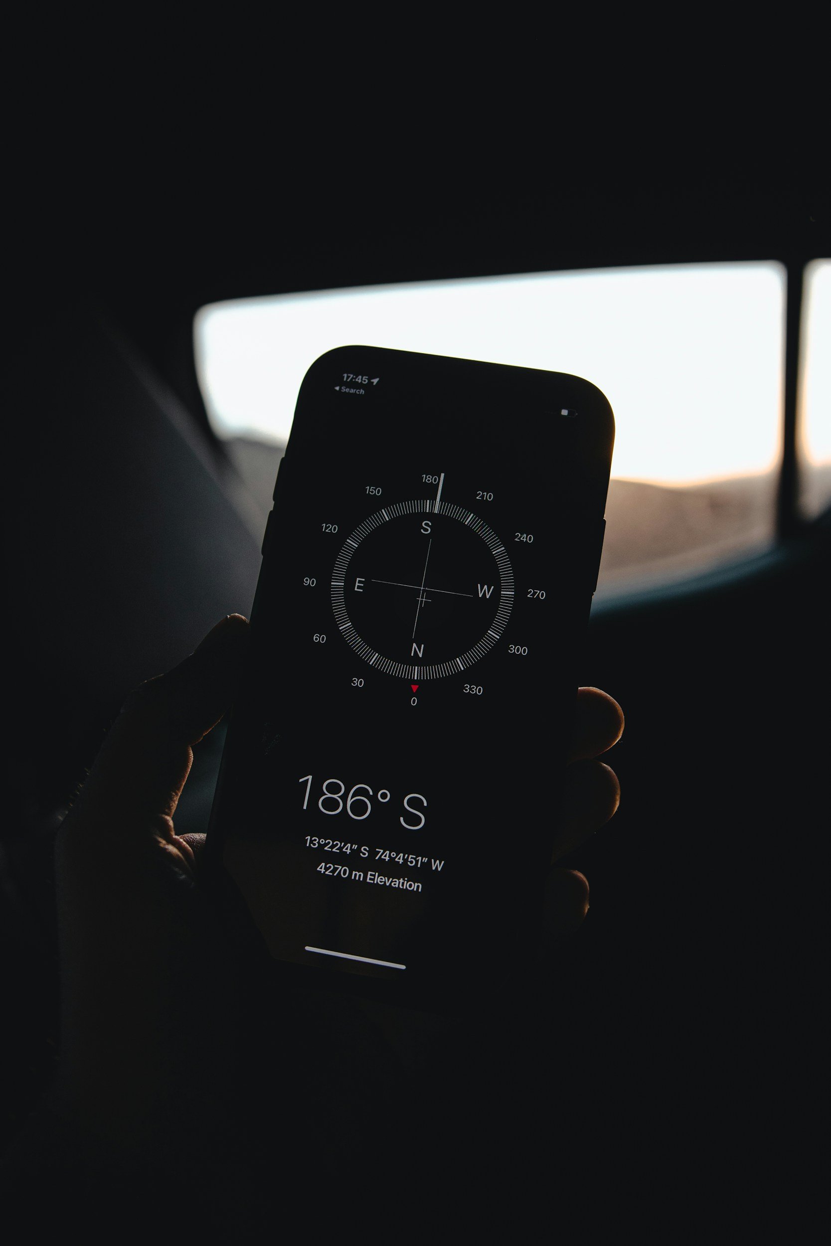 A person holding a smartphone displaying a compass app with a heading of 186 degrees south, with coordinates 13°22'44"S 74°4'51"W and an elevation of 4270 meters, inside a vehicle during sunset or sunrise.