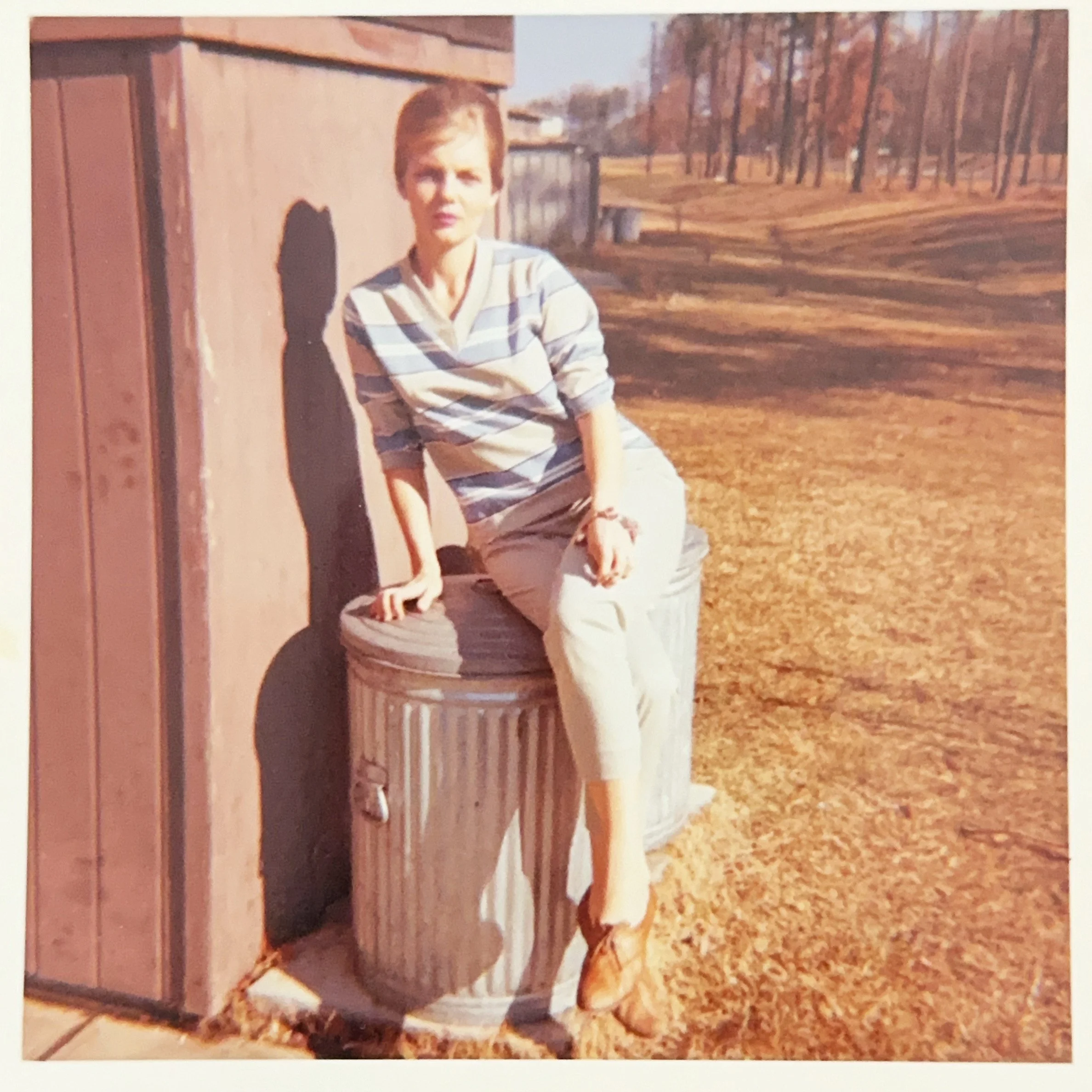 A woman with short hair wearing a striped long-sleeve shirt, light-colored pants, and brown shoes, sitting on a large trash can outdoors in a park during autumn or winter with leafless trees in the background.