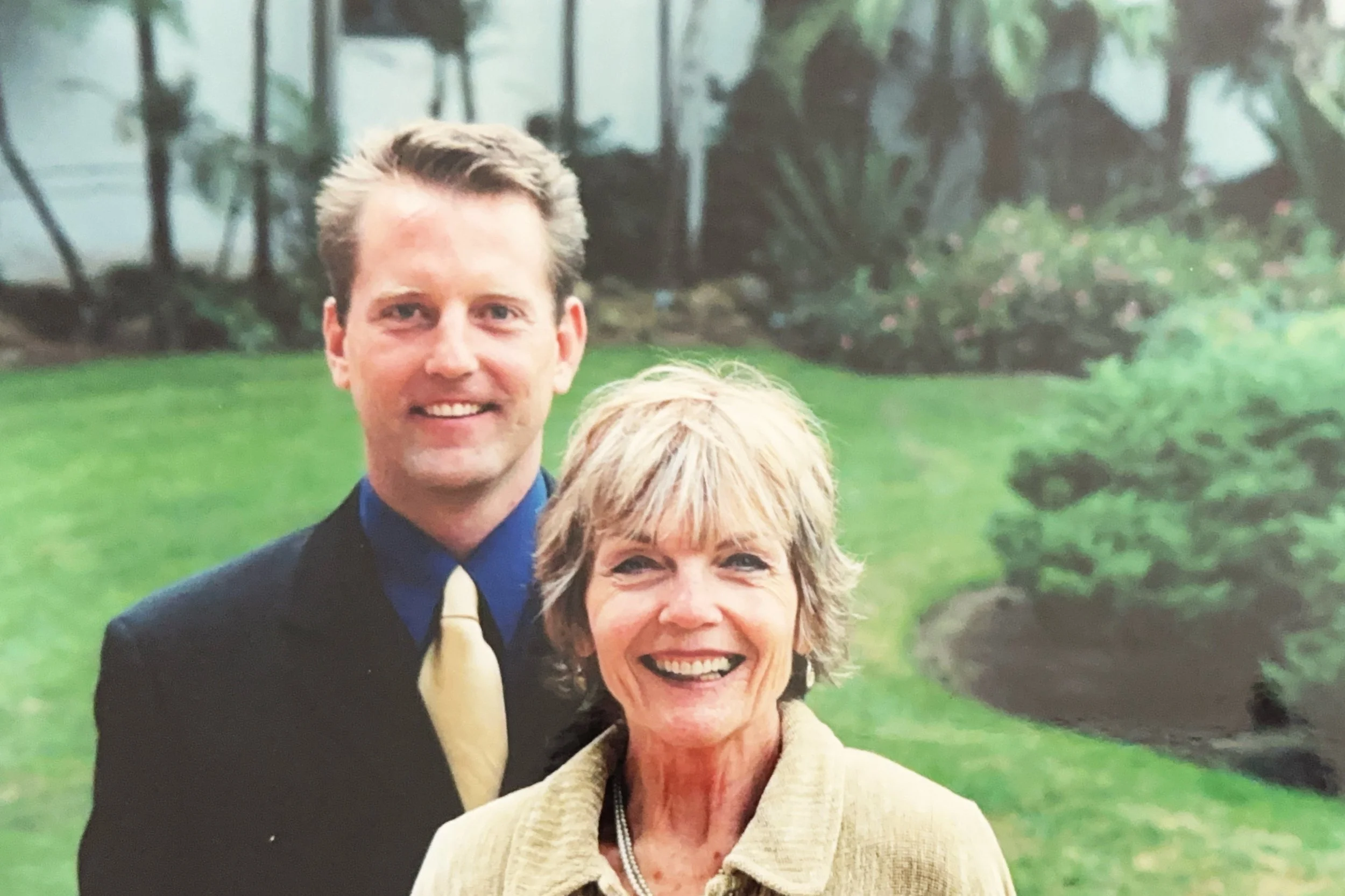 A young man and an older woman standing outdoors on a lawn with greenery in the background, both smiling at the camera.