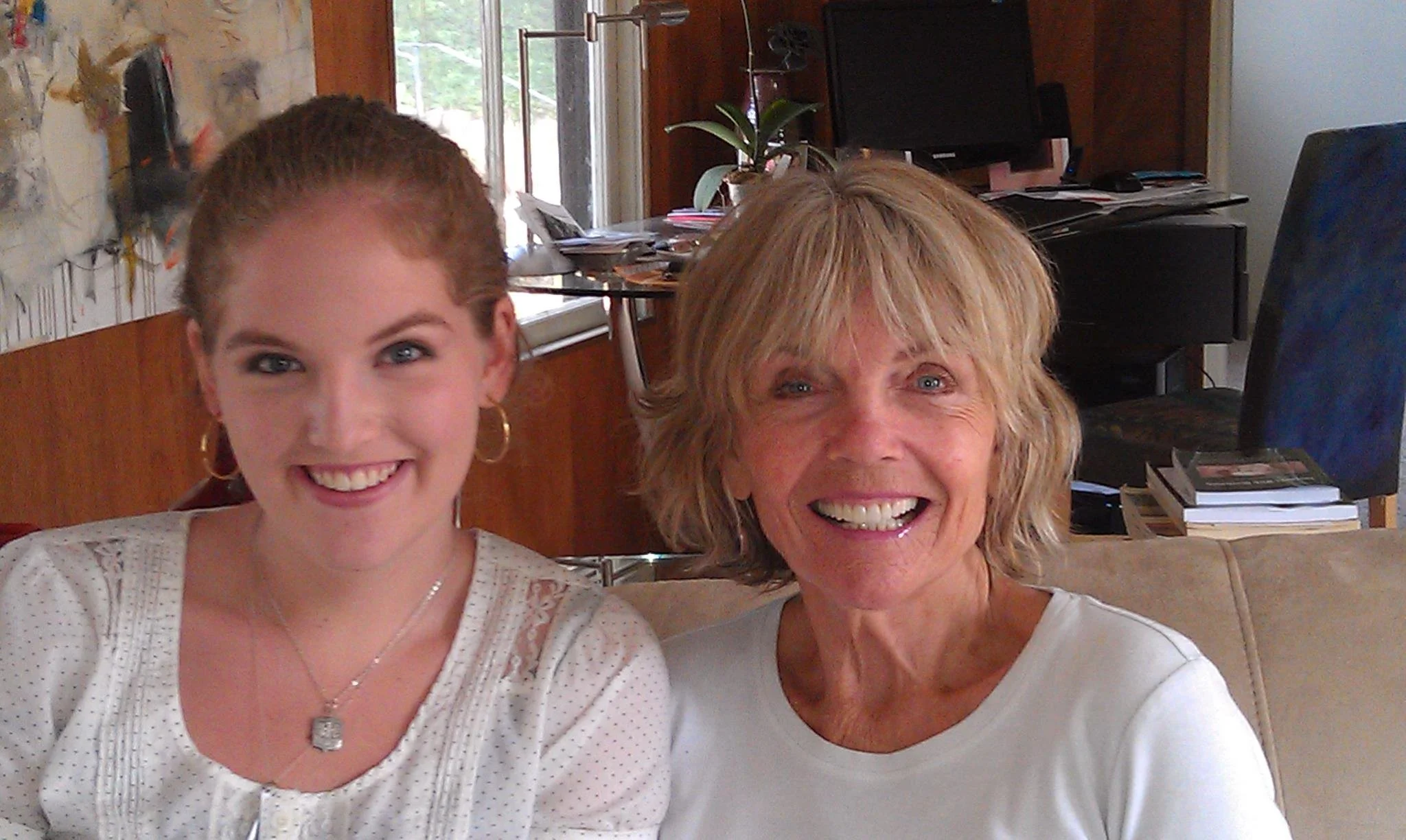 A young woman with light skin, brown hair tied back, wearing a white top with lace details, and jewelry, sitting next to an older woman with light skin, blonde hair, wearing a white t-shirt, both smiling inside a room with a couch, TV, computer, and cluttered desk in the background.