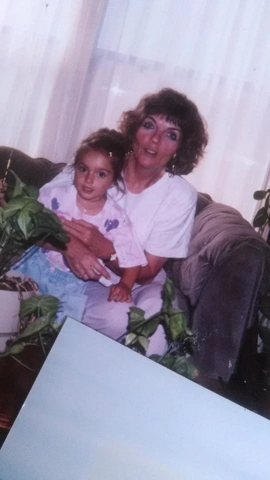 An older woman with curly hair sitting on a couch holding a young girl with a bow in her hair. There are houseplants and a white piece of paper on a table in the foreground.