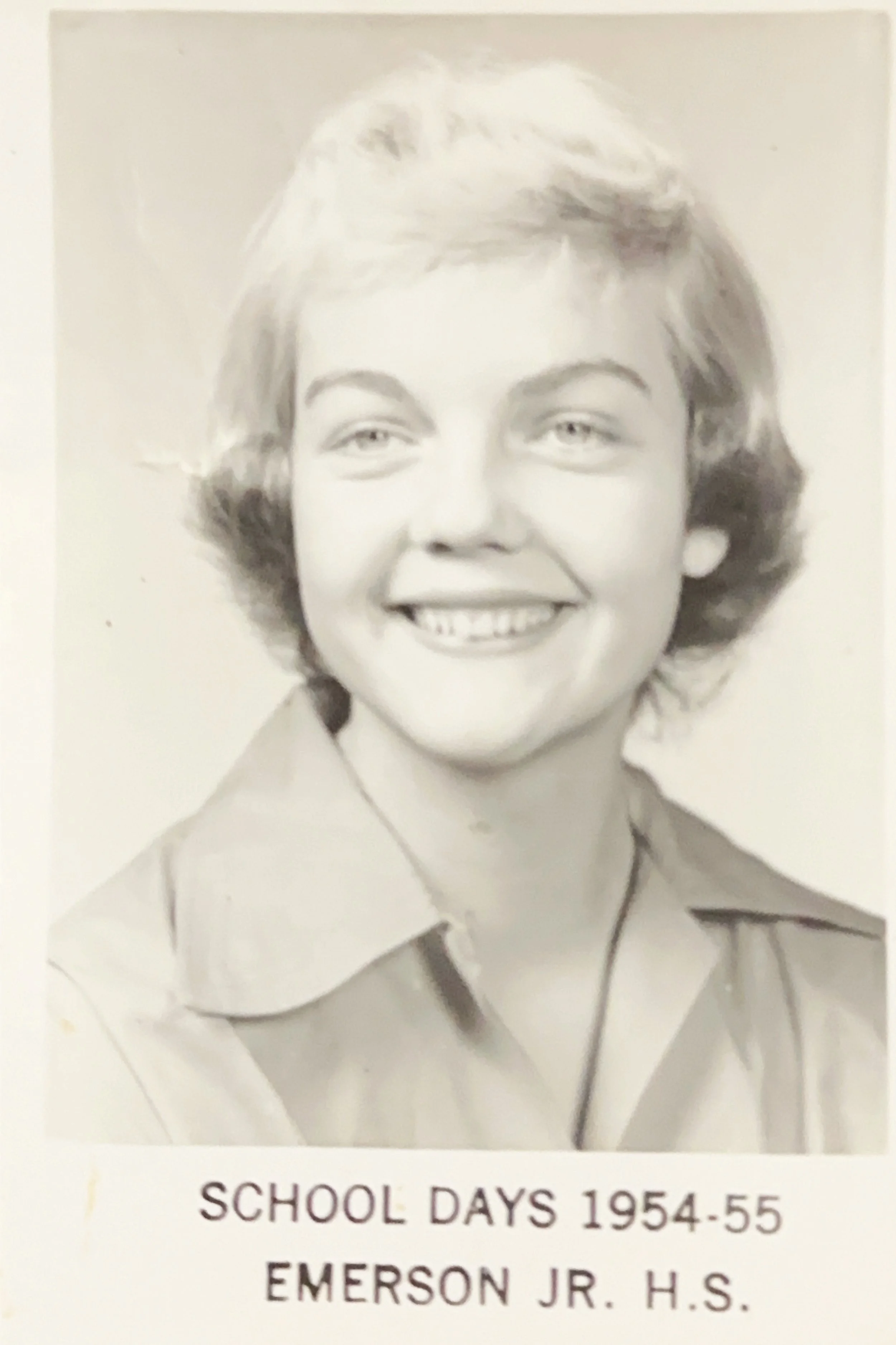 A black-and-white photo of a woman with short, wavy hair, wearing a collared shirt. Text at the bottom reads "School Days 1954-55, Emerson Jr. H.S.".