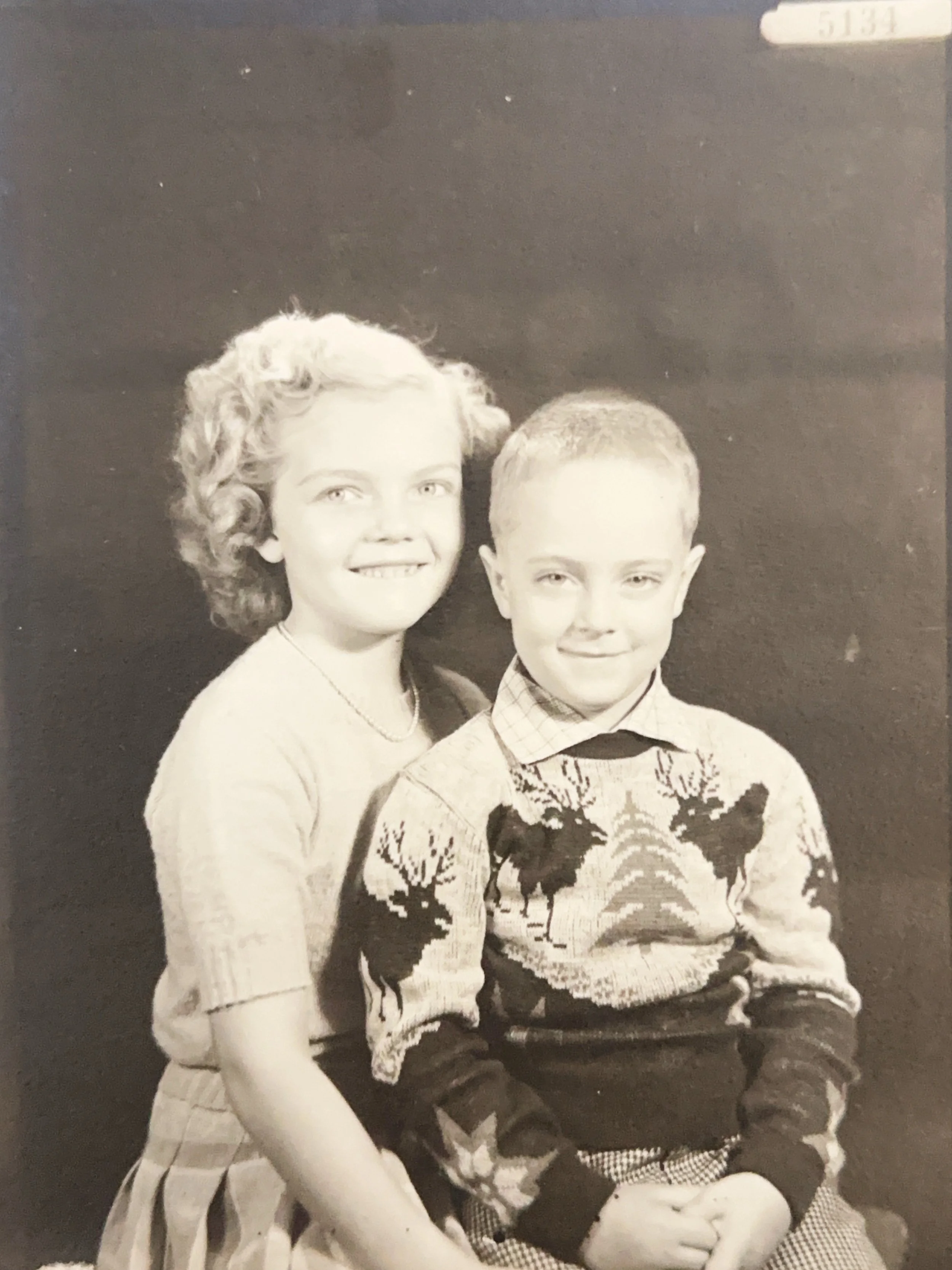 A black and white photograph of a young girl with curly hair and a young boy with short hair, both smiling and sitting close together against a plain dark background.