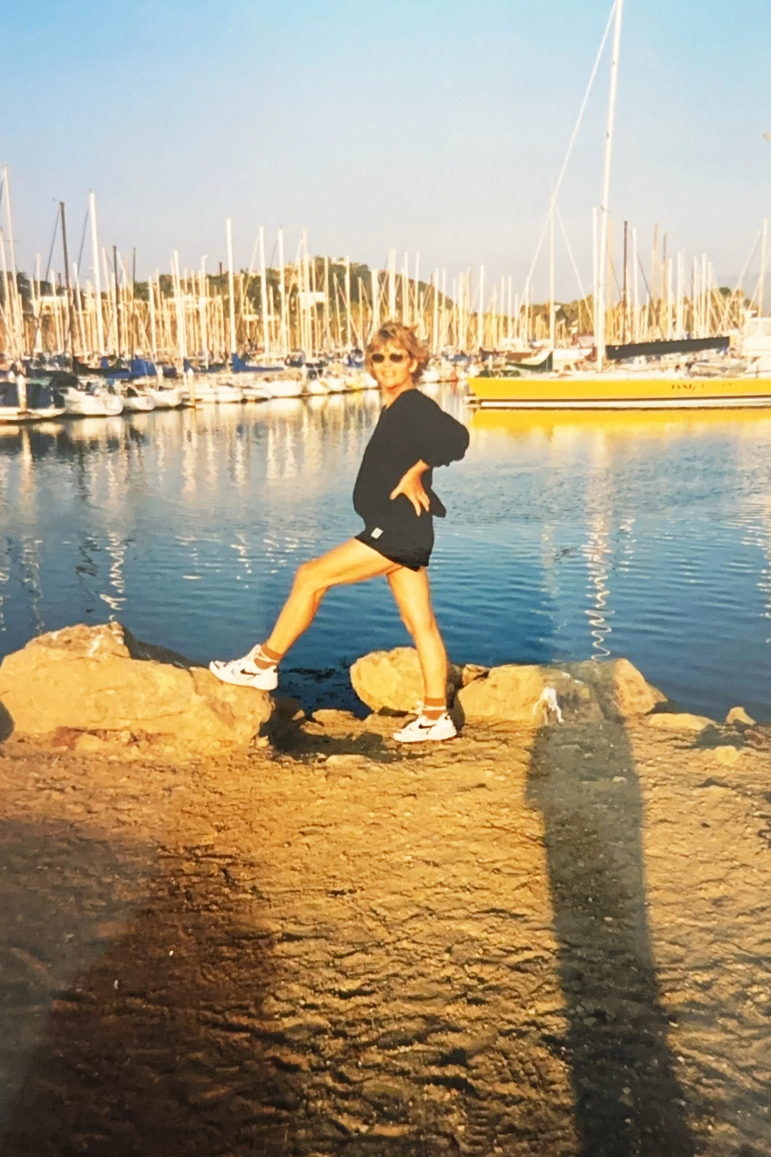 A woman standing on rocks by a marina with boats, wearing a black hoodie, shorts, and sneakers during sunset.