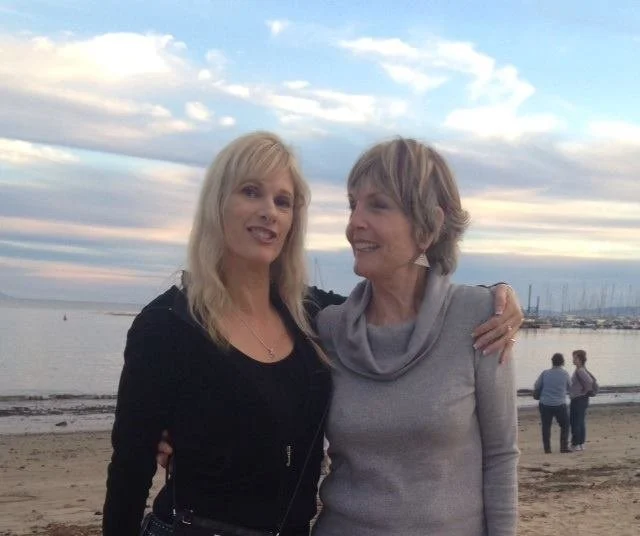 Two women standing on a beach during sunset, smiling and embracing each other, with a marina and boats in the background.