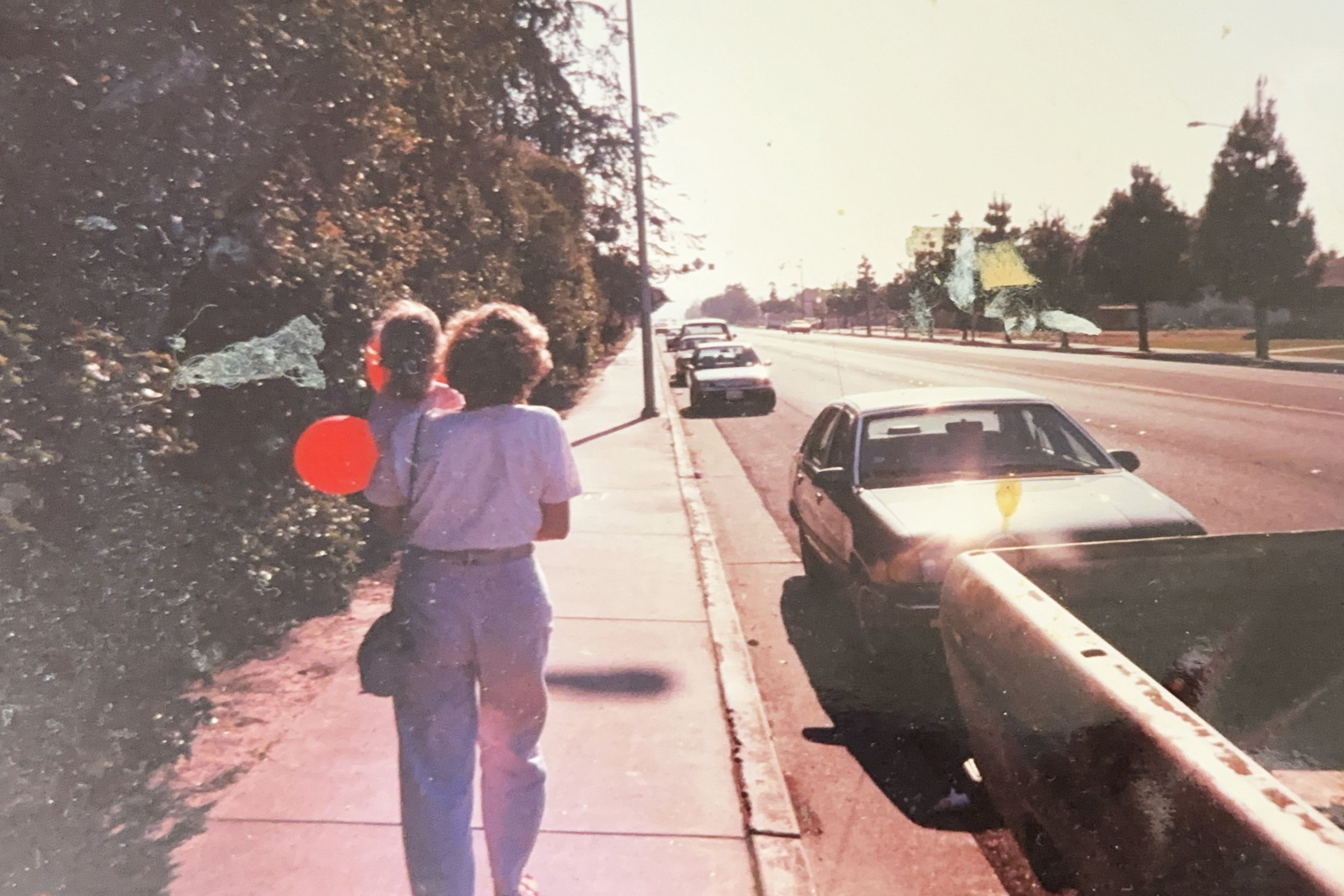 Two children, one holding a red balloon, walk on the sidewalk on a sunny day along a street with parked cars and trees in the background.