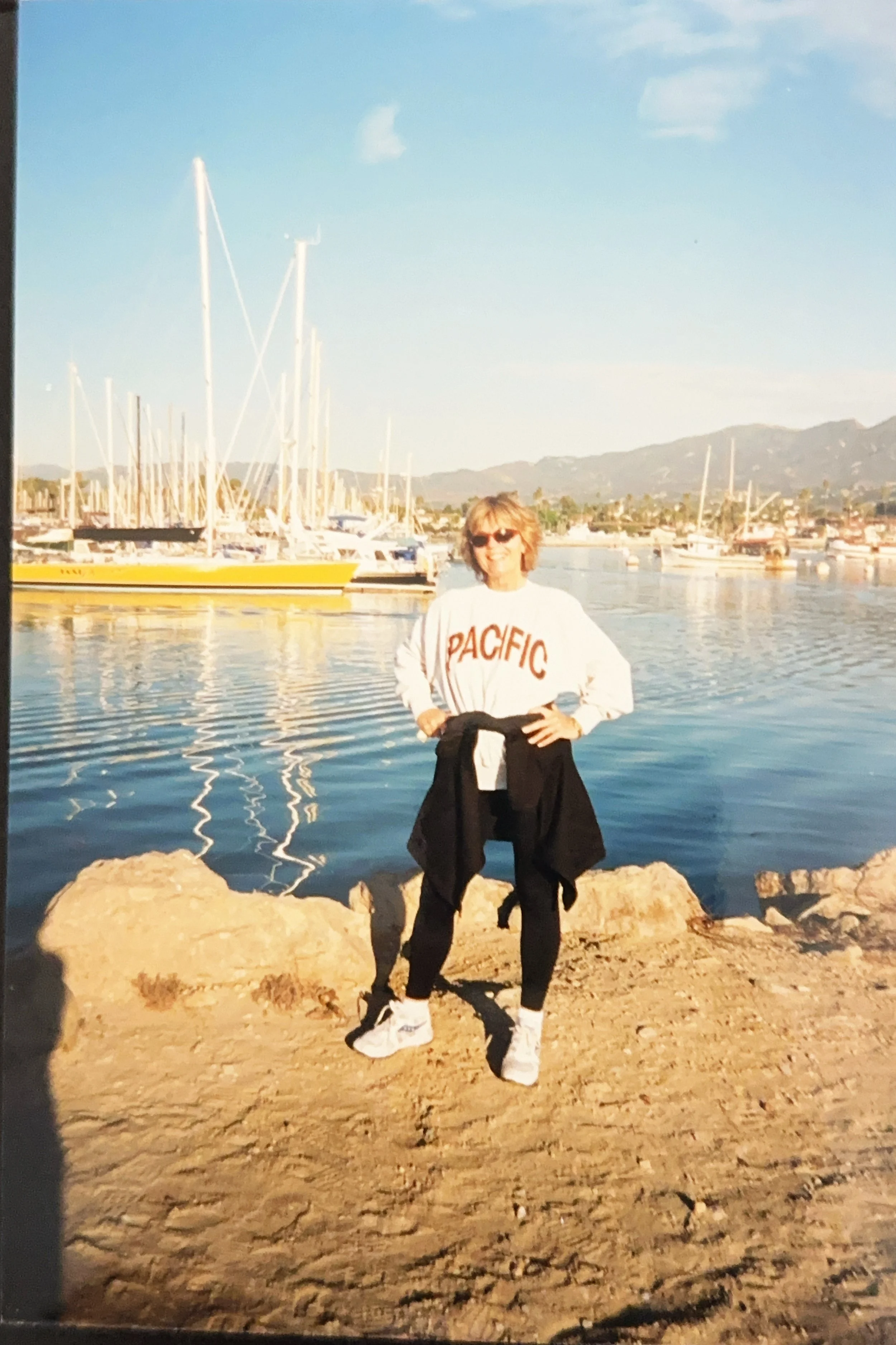 A woman standing on a rocky shore near a marina with sailboats docked and mountains in the background, wearing a white sweatshirt with 'PACIFIC' written on it, sunglasses, and athletic clothing.