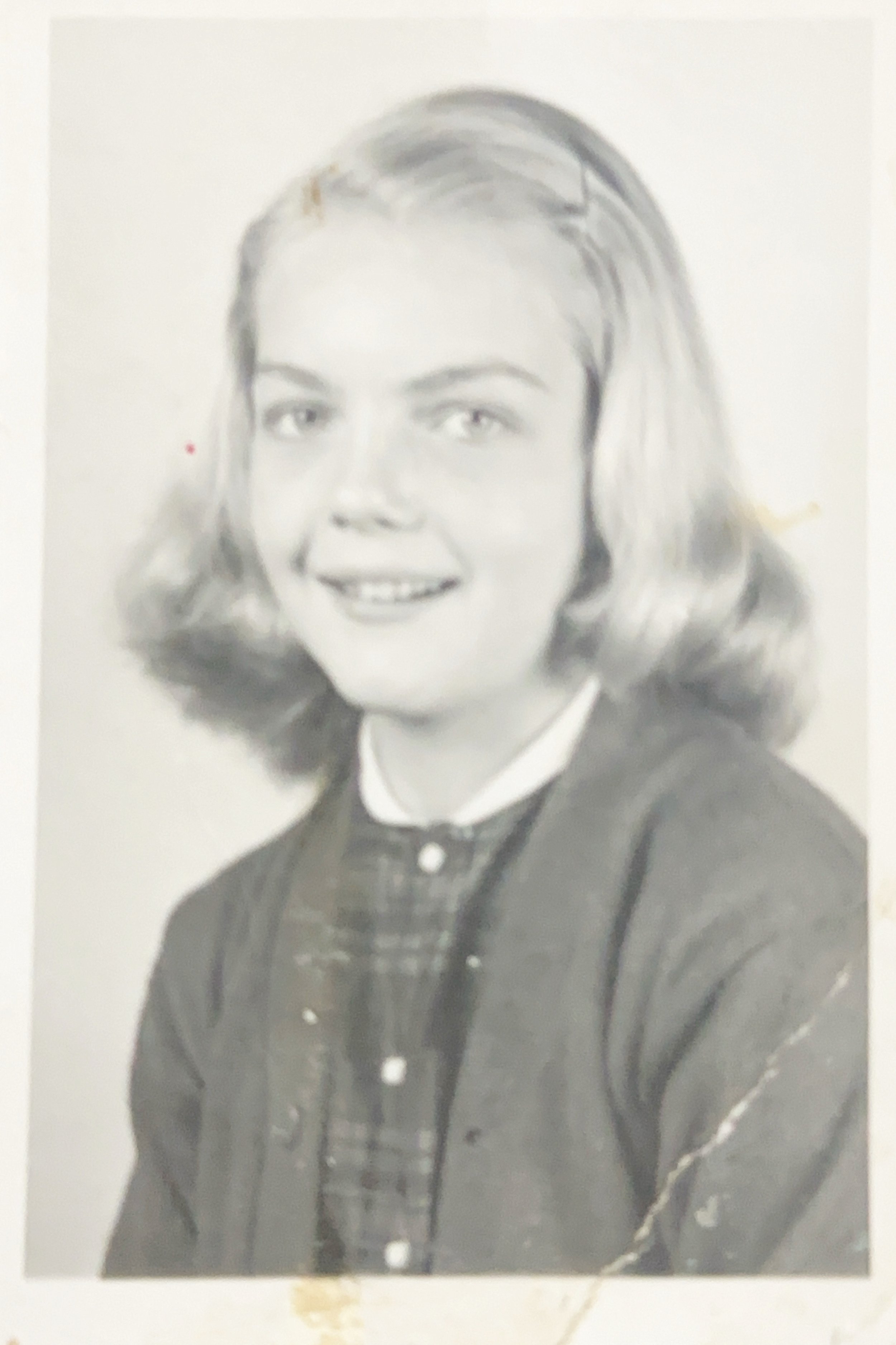 A black and white photograph of a young girl with shoulder-length wavy hair, wearing a plaid shirt and a cardigan, smiling at the camera.