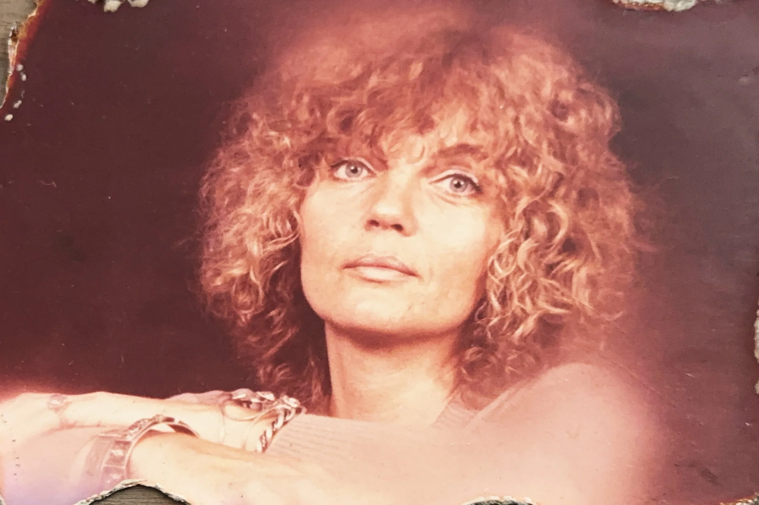 A woman with curly, reddish-blonde hair and light eyes looking directly at the camera, resting her arm on a surface.