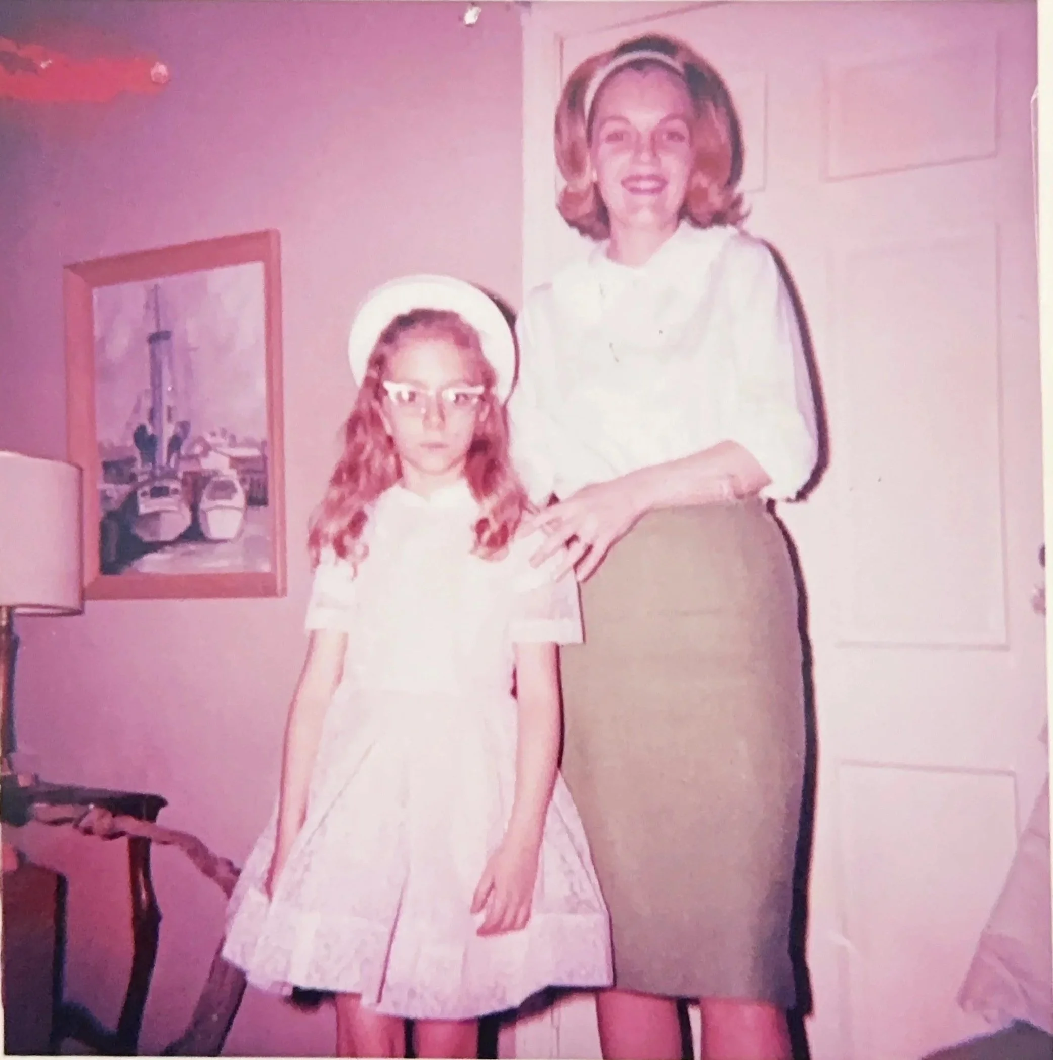 A woman with blonde hair and a girl with curly red hair are posing together in a room. The woman is standing behind the girl with one hand resting on her shoulder. The girl is wearing glasses and a white dress with a full skirt, and the woman is wearing a white top and a brown skirt. There is a framed picture on the wall and a table lamp in the background.
