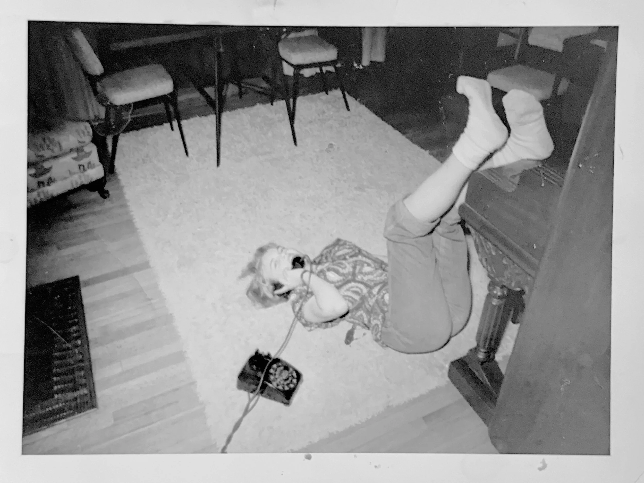 A child lying on a rug in a room, talking on a vintage rotary phone, with their legs resting on a piano. The room has wooden floors, a small sofa, and chairs.