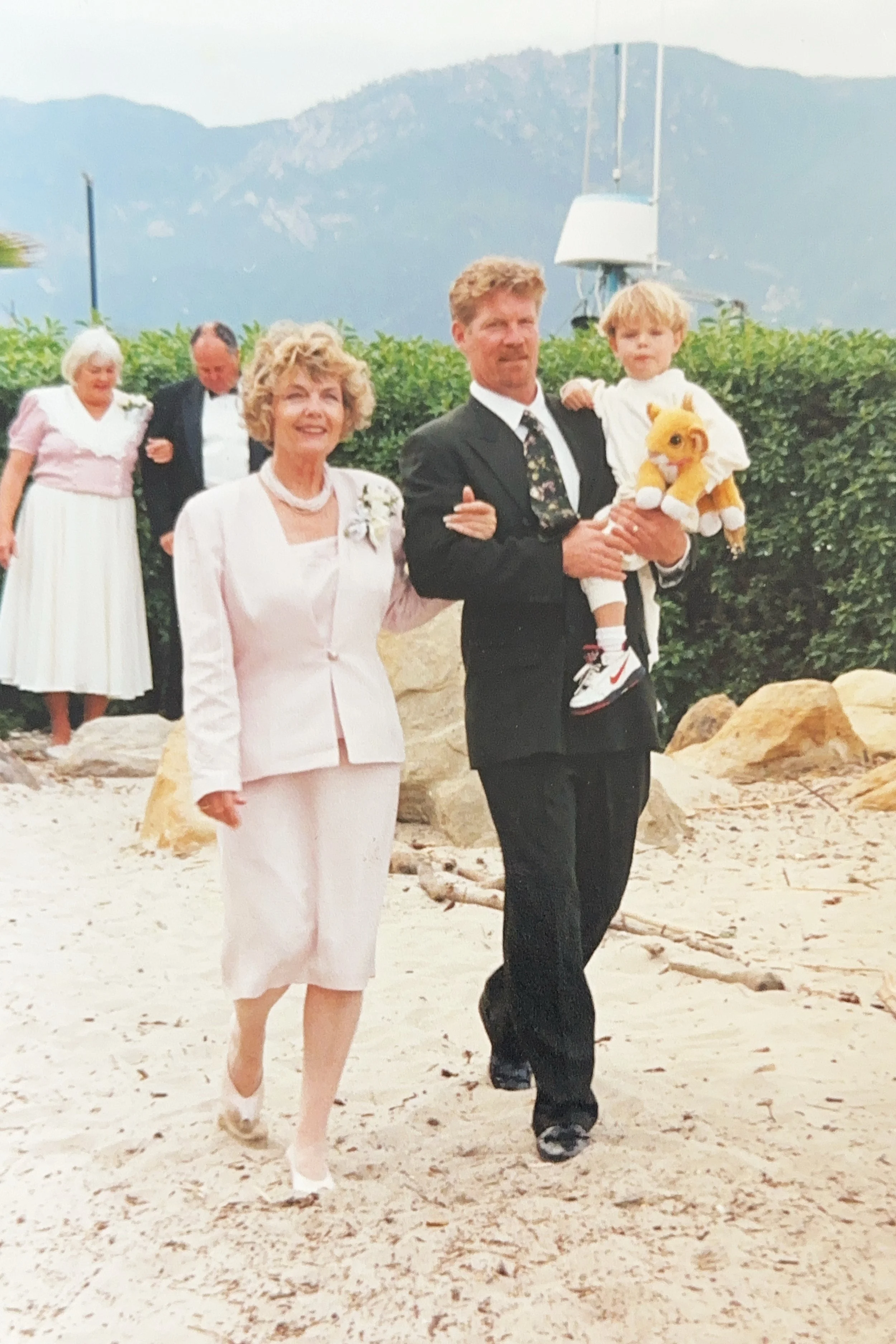 A man in a black suit carrying a young boy in a white outfit and holding a plush lion, walking on a sandy beach with a woman in a light pink suit walking beside him. In the background, two elderly people walk together near greenery, with mountains and a cloudy sky in the distance.