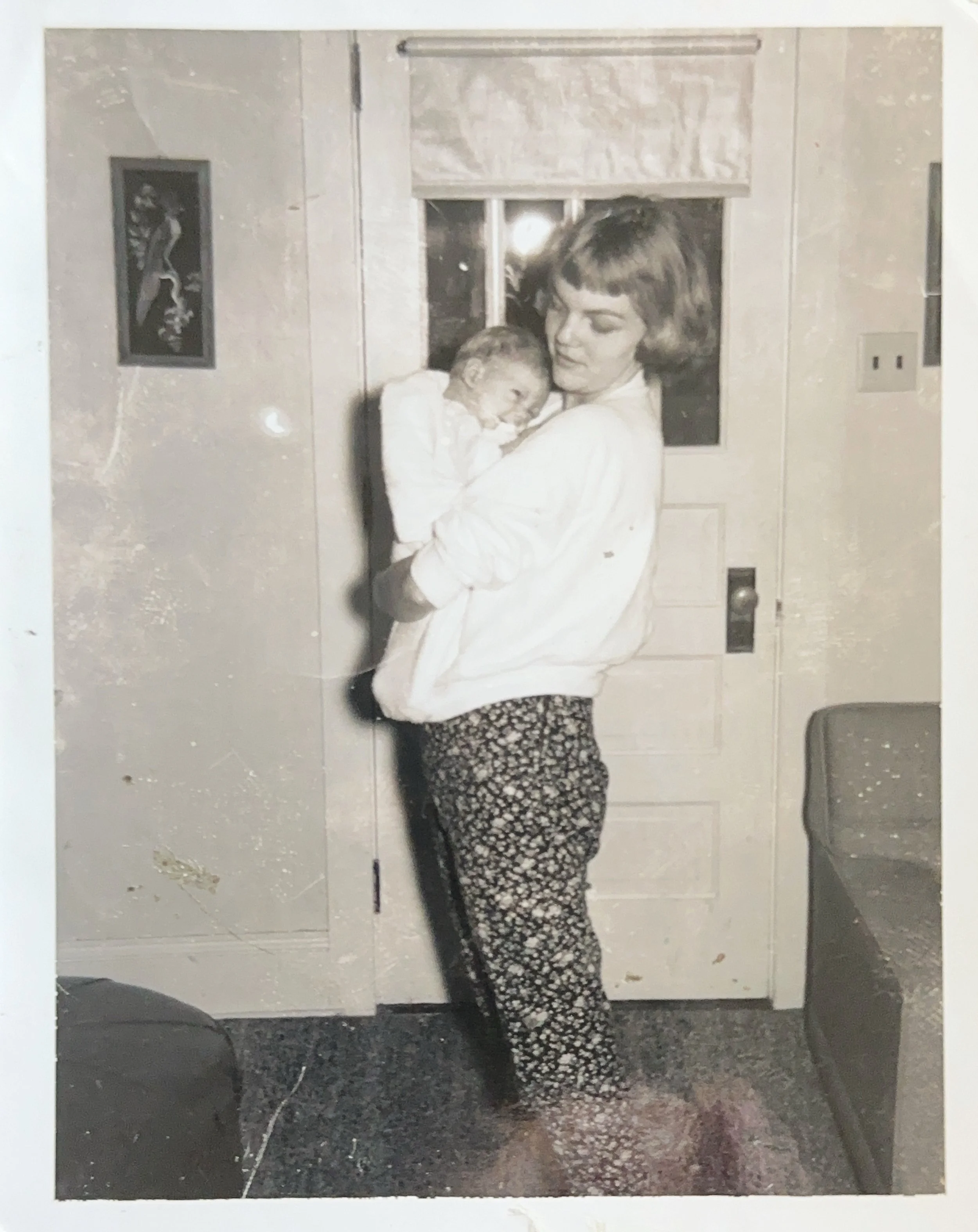 A woman holding a baby inside a house near a door with a window and a picture on the wall
