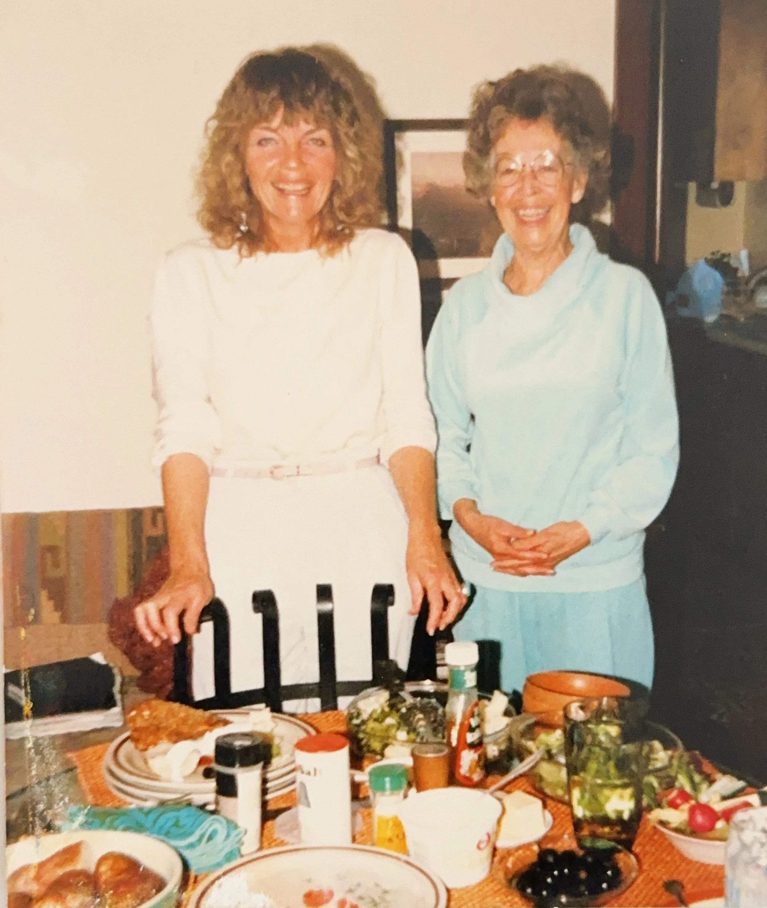Two women smiling and standing behind a table with food, condiments, and bowls, in a home kitchen or dining area.