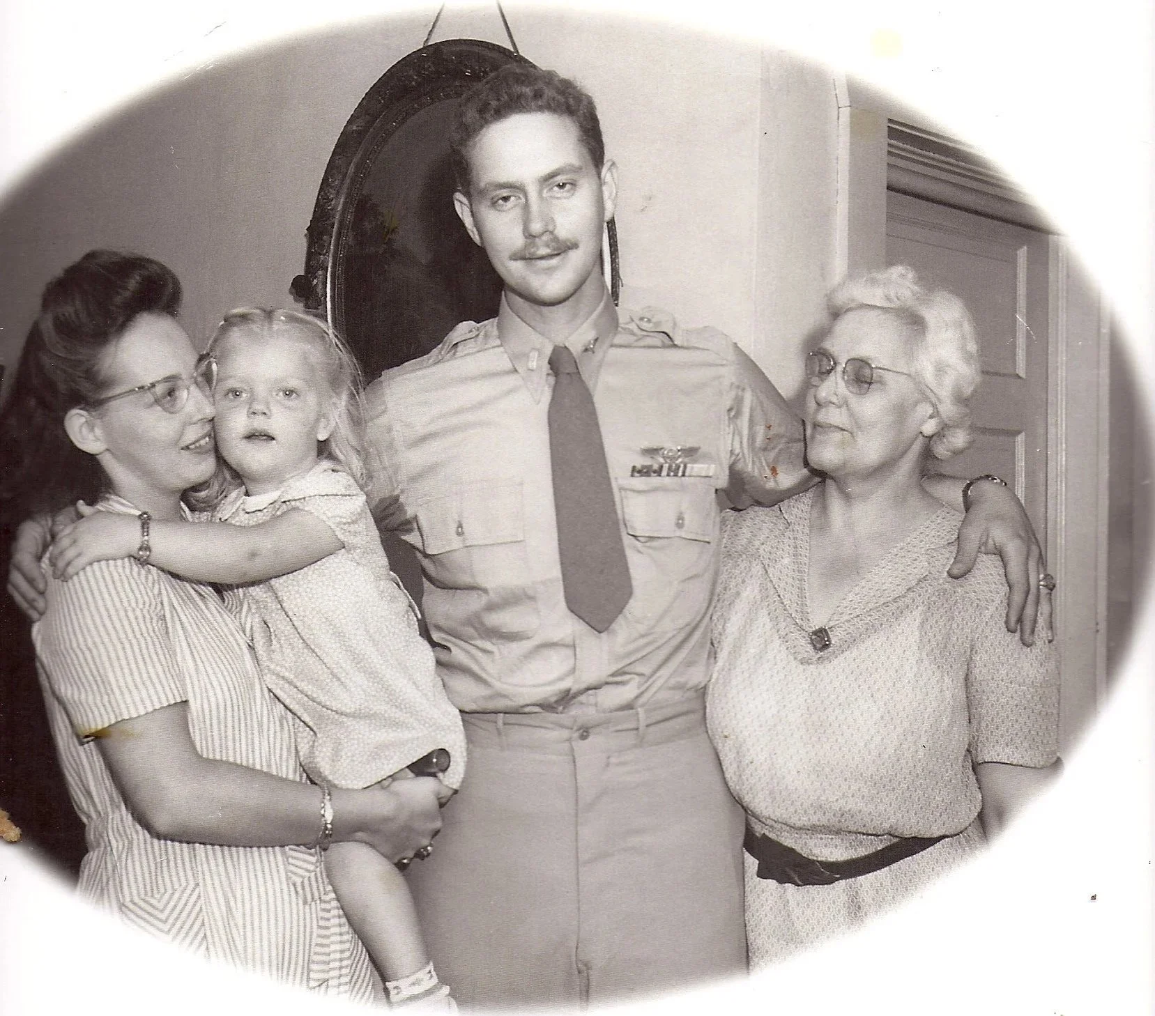 Black and white photo of four people, including a woman holding a young girl, a man in a military uniform, and an older woman, standing close together and smiling.