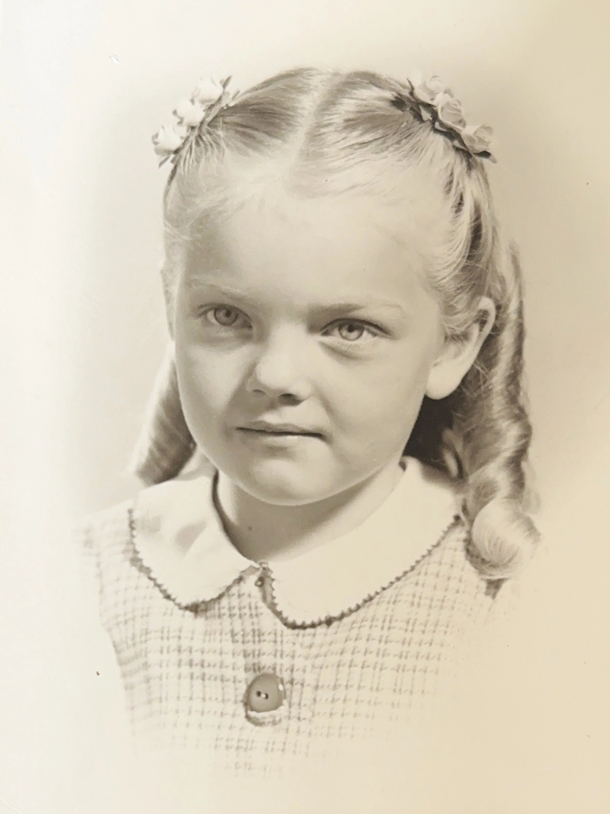 Black and white portrait of a young girl with curly hair and light-colored eyes, wearing a gingham dress with a Peter Pan collar and a large button, looking slightly to the side.