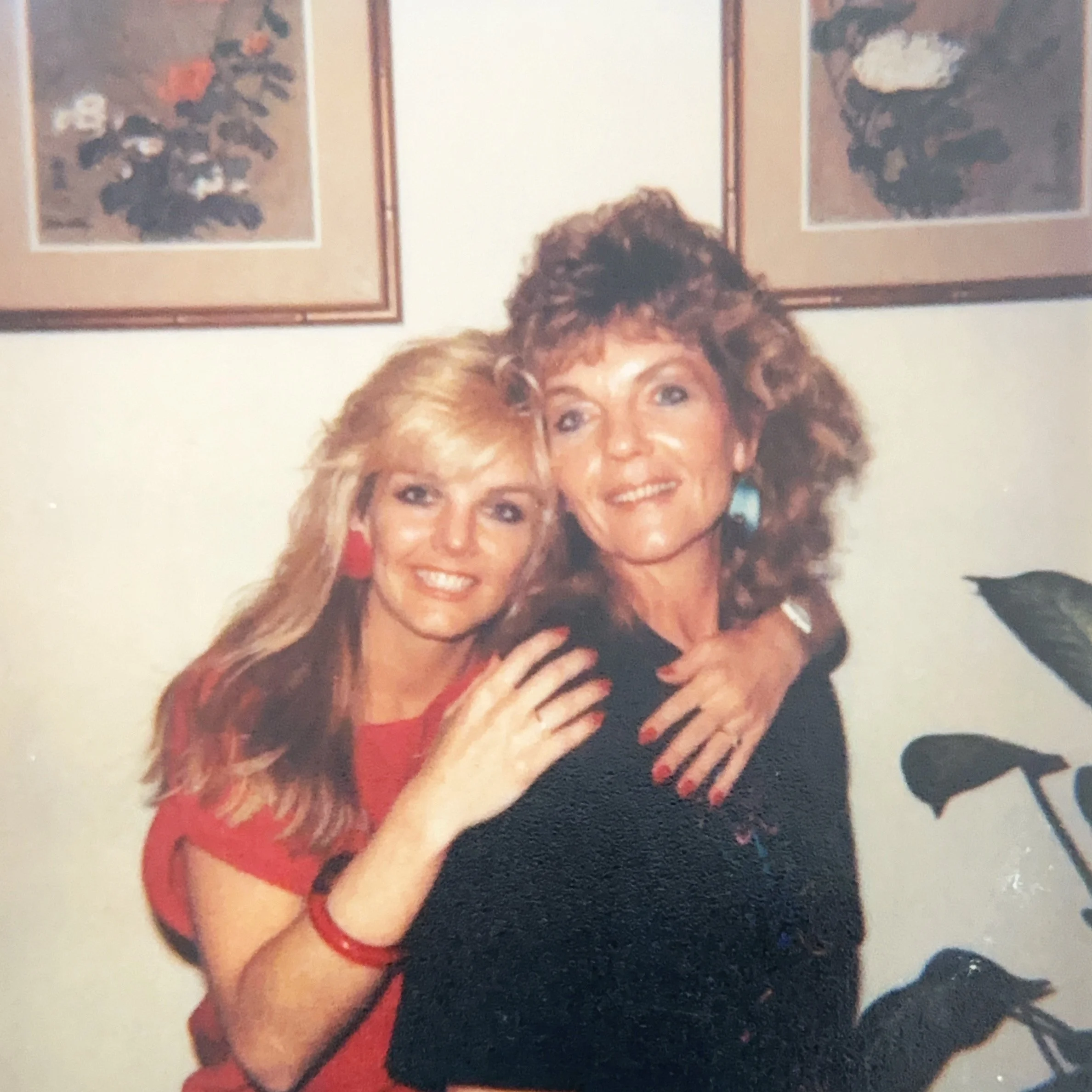 Two women hugging, smiling indoors with framed floral art on the wall behind them and a potted plant to the side.