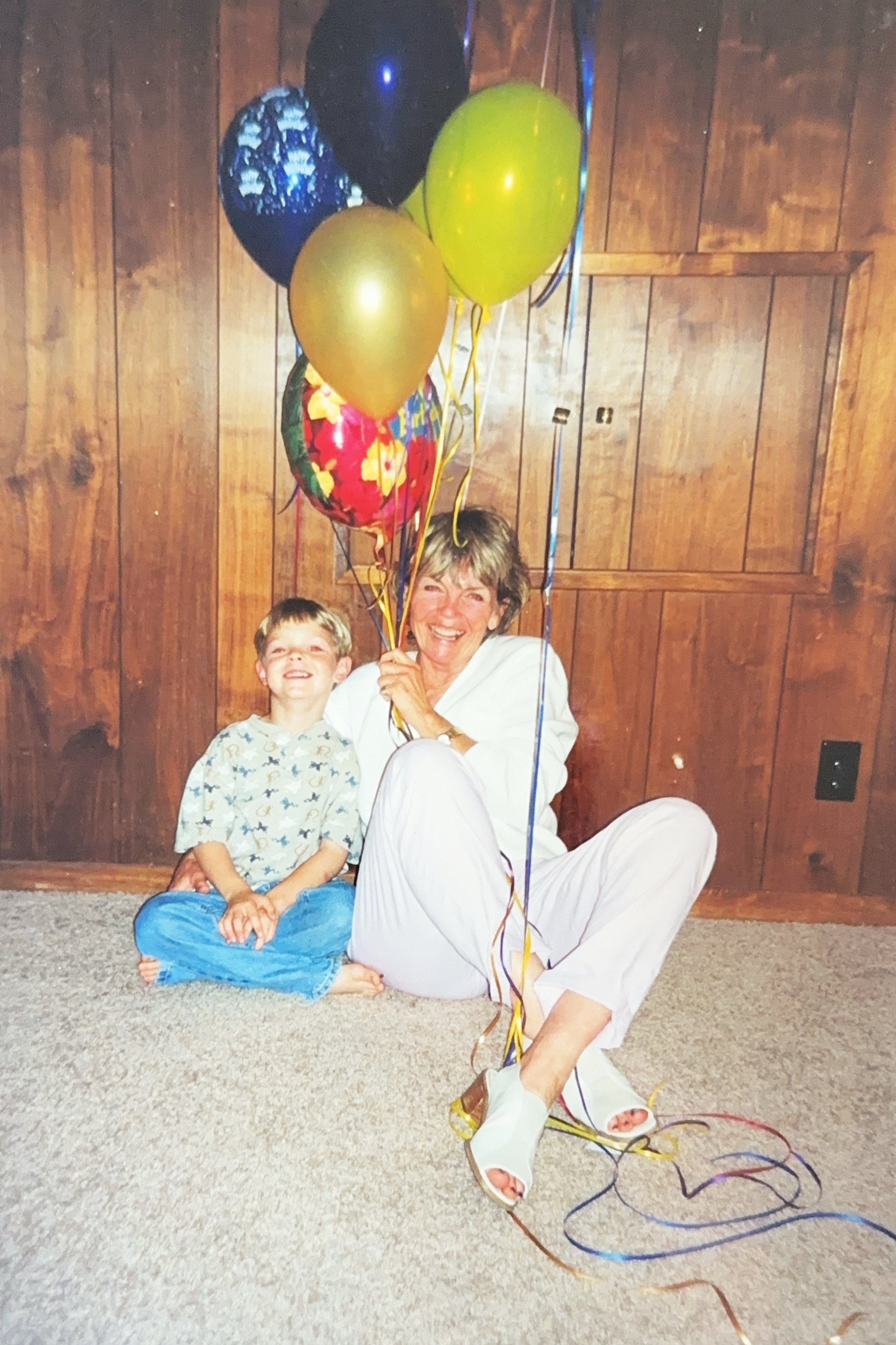 A woman and a young boy sitting on a beige carpet, smiling and holding a bunch of colorful balloons against a wooden-paneled wall.