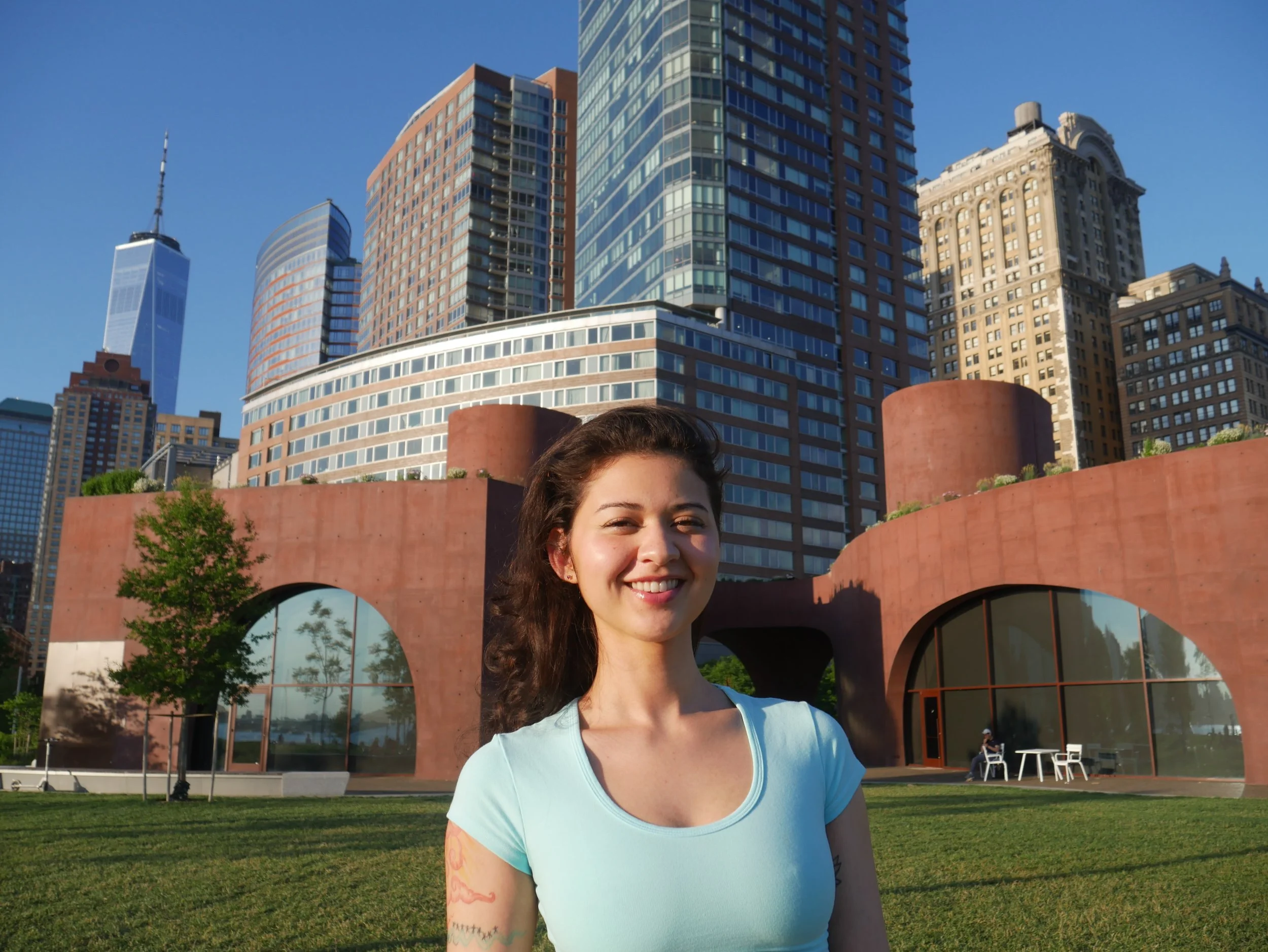 Smiling woman with dark, wavy hair standing in front of a city skyline featuring tall modern buildings, a green park, and a clear blue sky.
