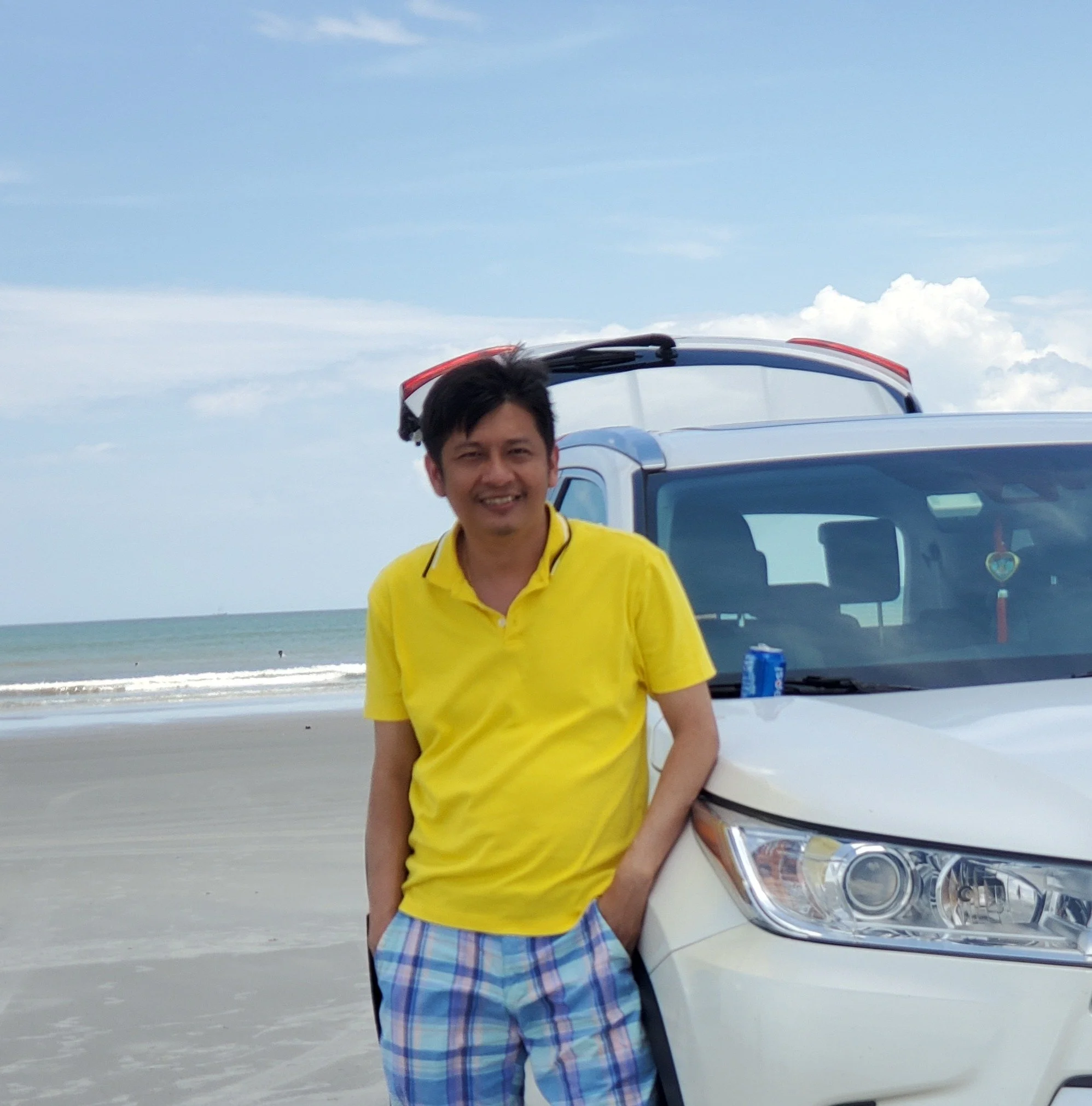 A man in a yellow shirt and plaid shorts leaning against a white SUV on a beach, with the ocean and blue sky in the background.