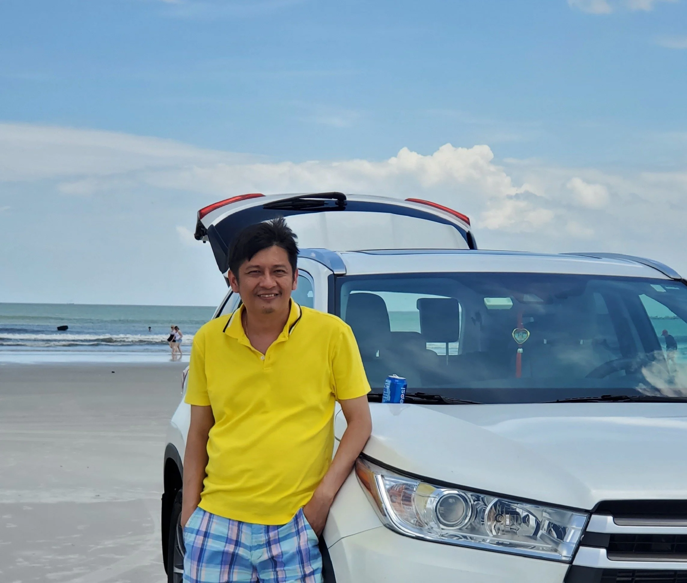A man in a yellow shirt and plaid shorts leaning against a white SUV on a beach with the ocean and blue sky in the background.