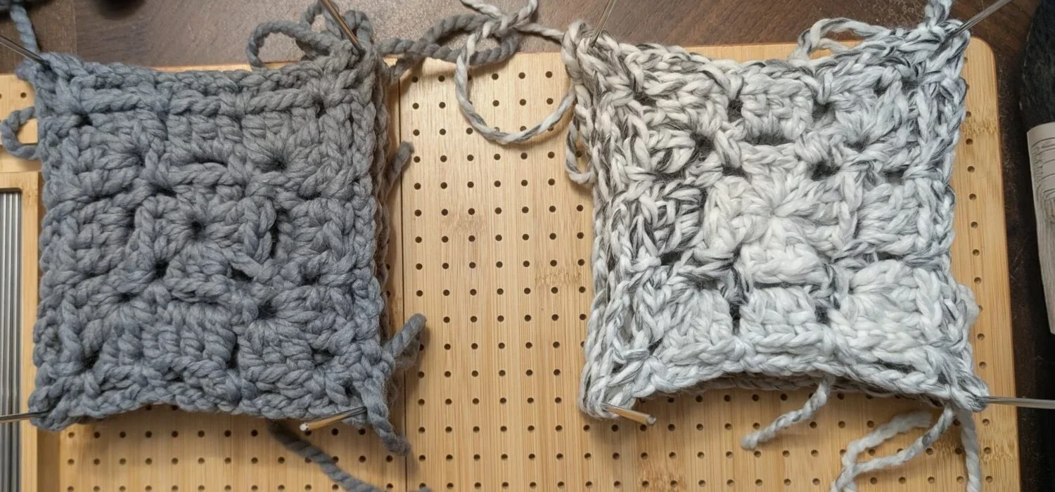 Two crocheted square swatches on a wooden knitting board with yarn and knitting needles, one in gray and one in white with gray streaks.