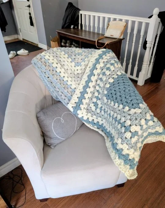 A cozy living room corner with a beige armchair draped with a multicolored crocheted blanket in shades of blue, cream, and gray. A gray pillow and a white yarn ball are on the armchair. In the background, there's a wooden table with a purse and some bags on it, a black coat hanging on the staircase railing, and a pair of white shoes near the front door.