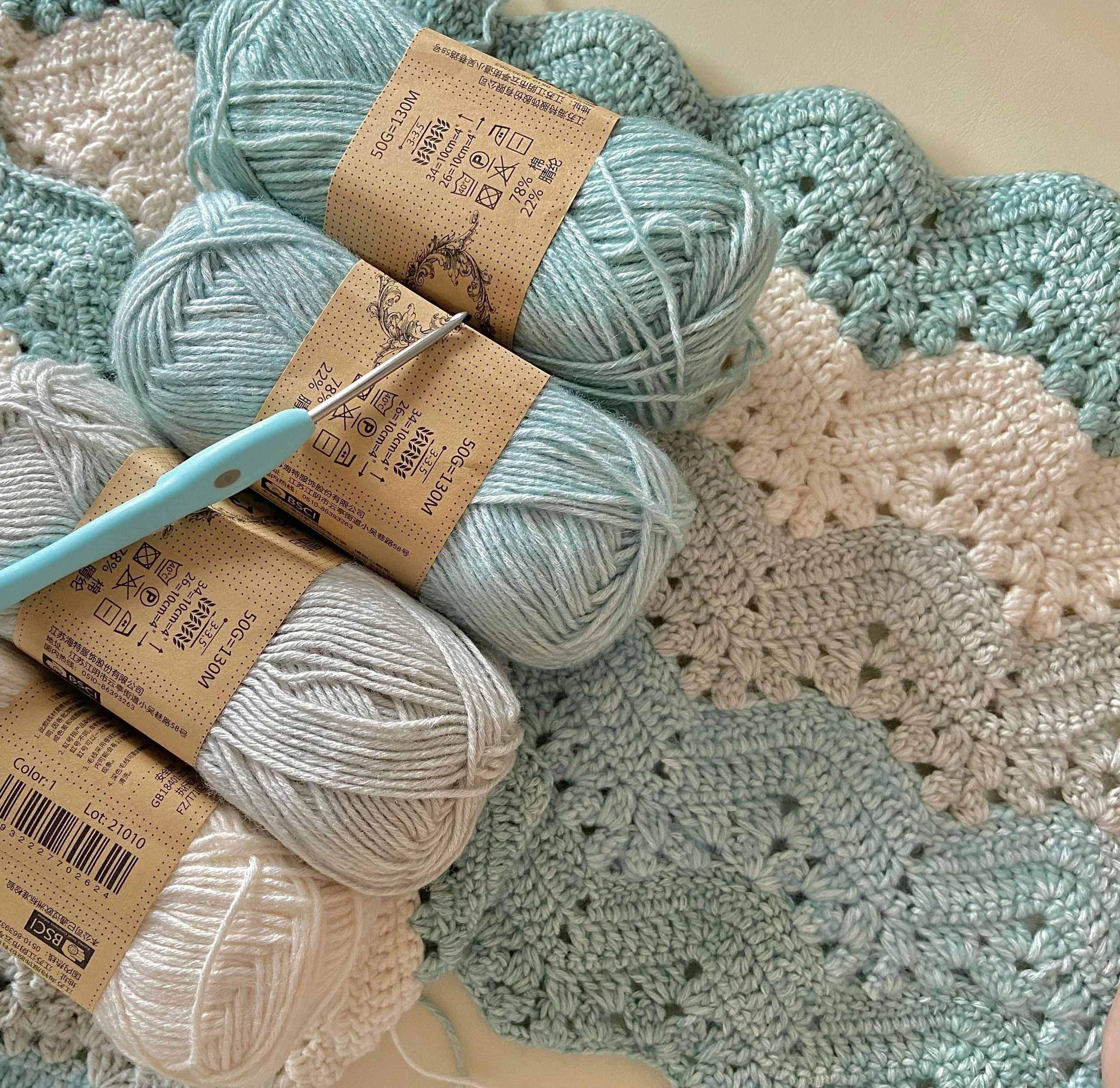 Several skeins of light blue and white yarn on top of a partially completed crochet blanket with blue, white, and light green squares.