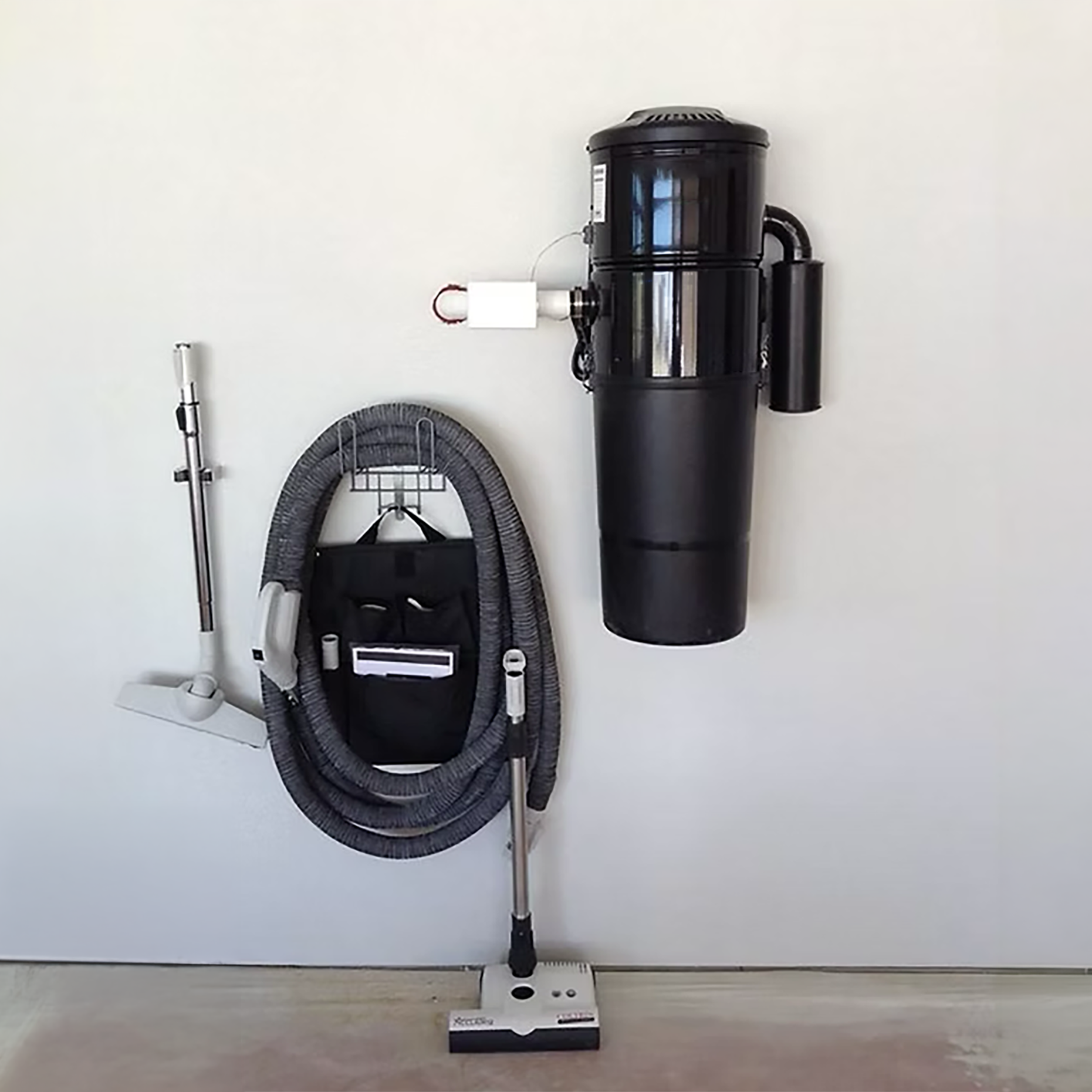A black robotic vacuum cleaner with a tall exhaust section, a coiled hose, and a disconnected metal wand, all mounted on a white wall and floor.