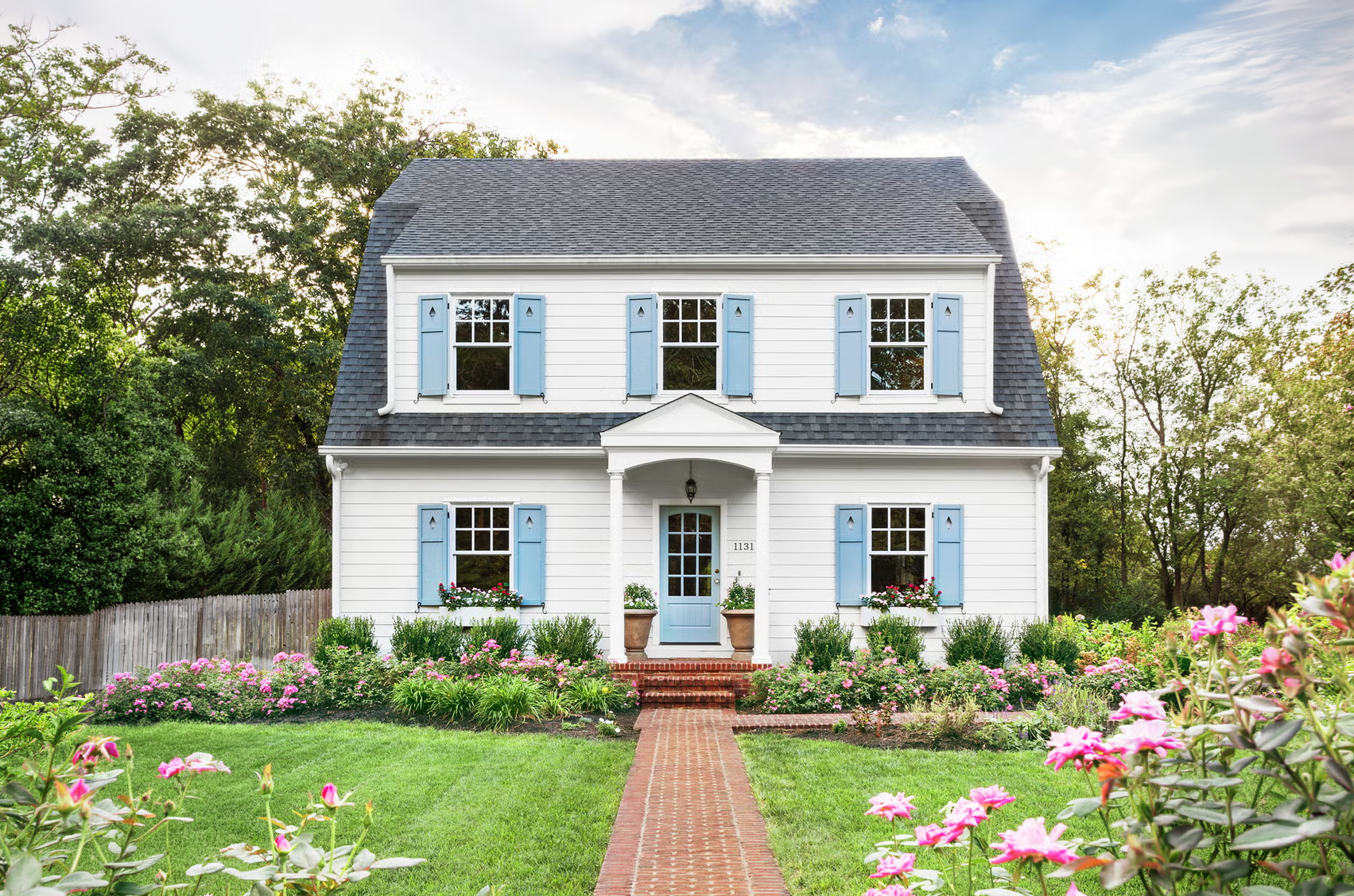 A white two-story house with light blue shutters and door, surrounded by a pink flower garden and green lawn, with trees in the background and a brick pathway leading to the front door.